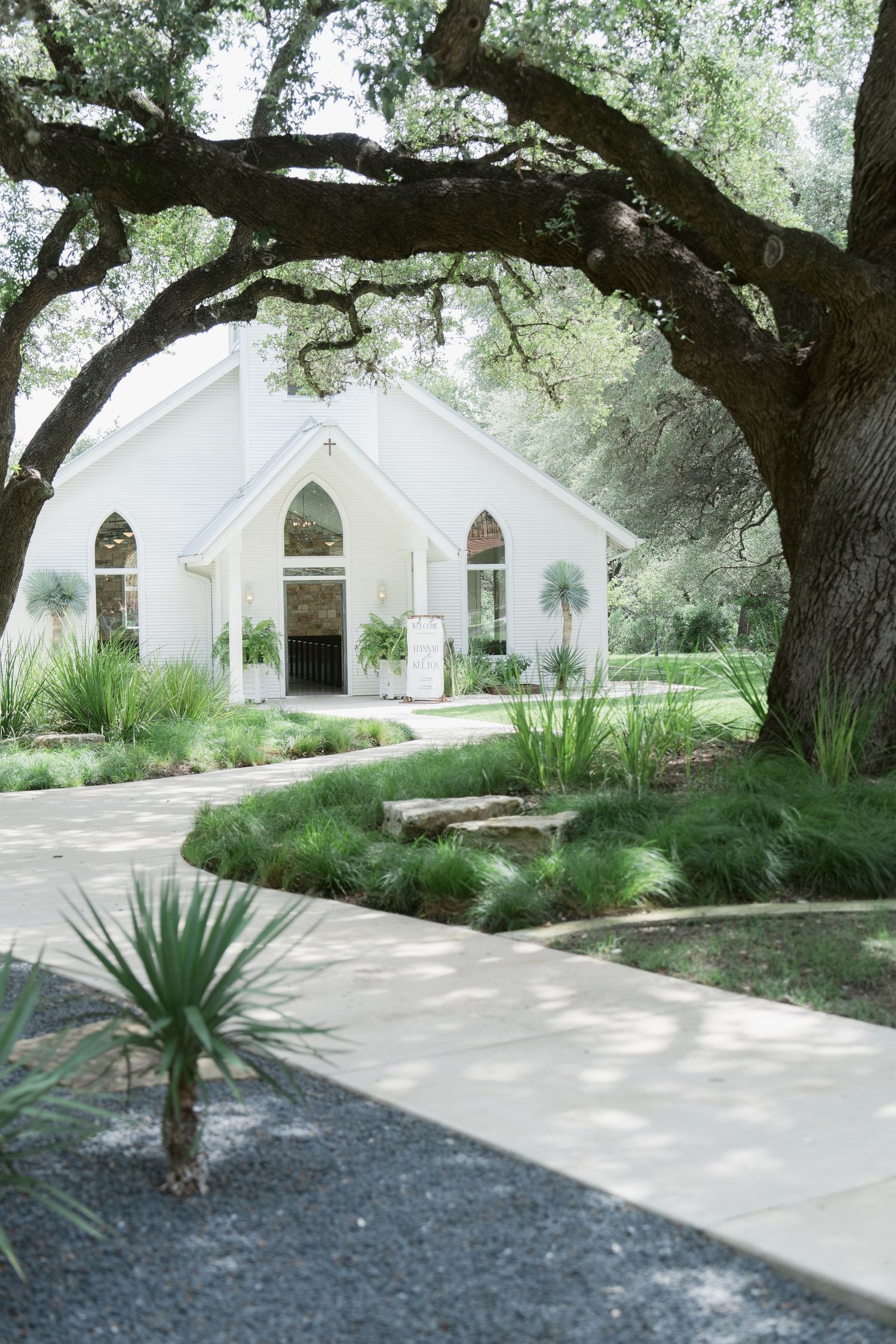White chapel surrounded by lush greenery and oak trees with a winding pathway leading to the entrance.