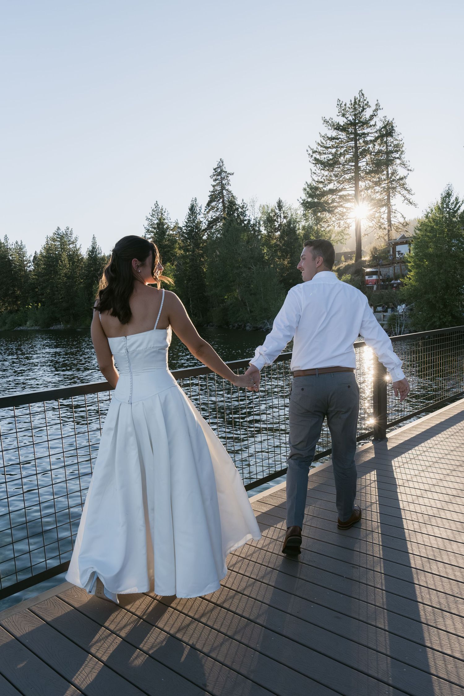 Bride and groom hold hands on a waterfront deck at sunset, surrounded by tall pine trees.