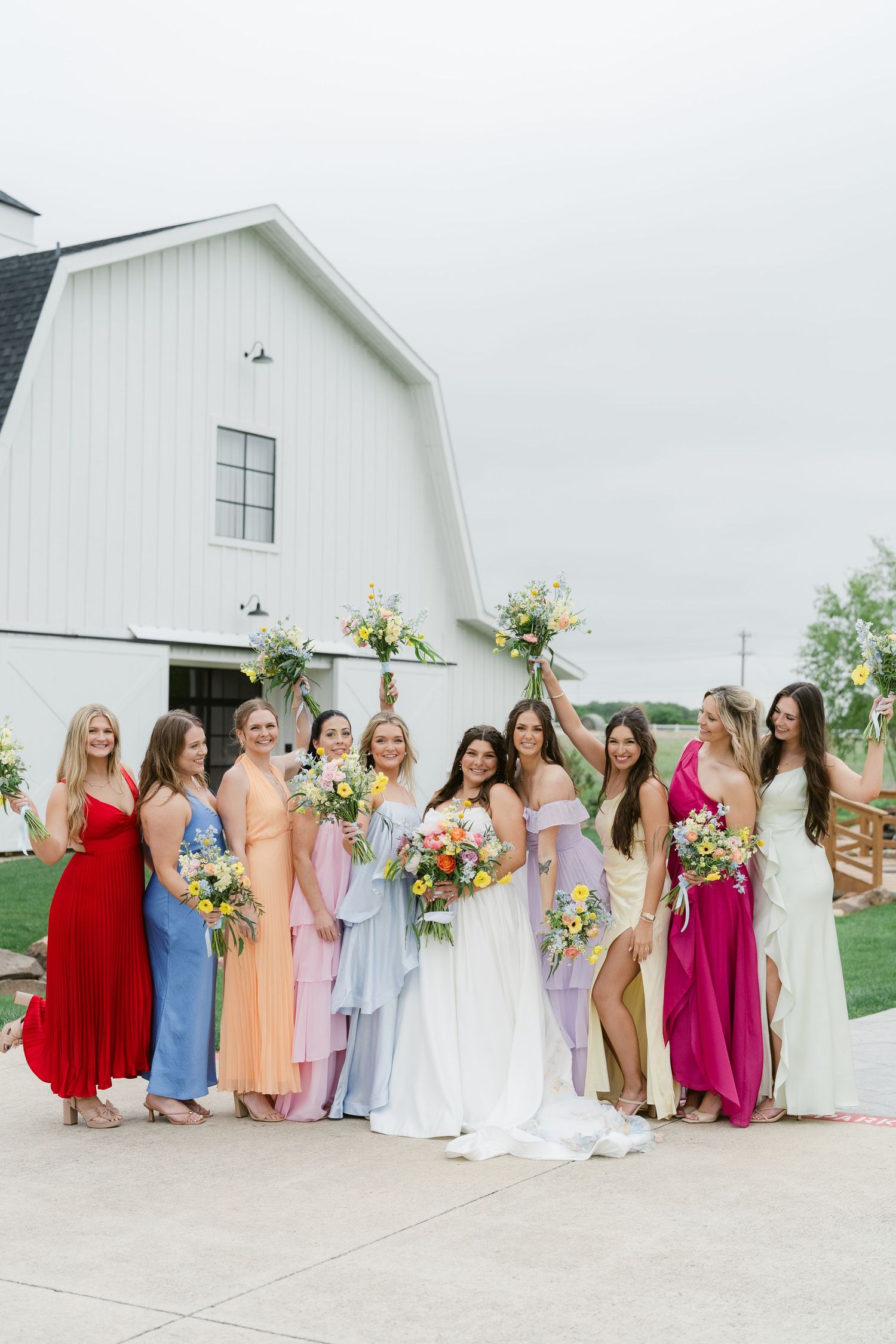 Bride in white gown poses with colorful bridesmaids holding floral bouquets outside a white barn venue.
