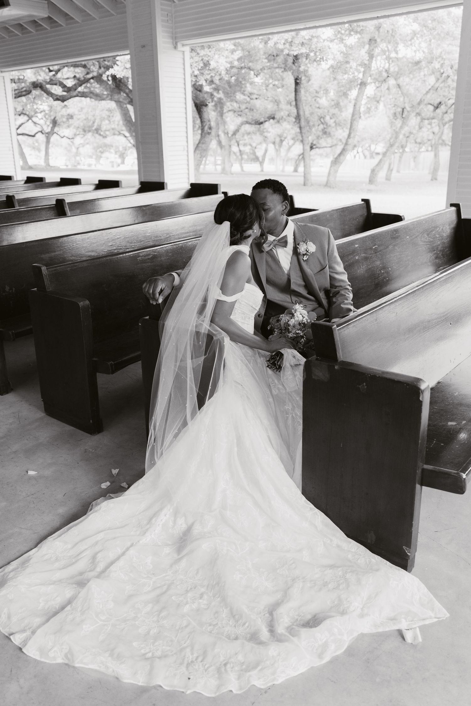 Black and white photo of a bride and groom sharing a kiss among wooden church pews, her veil and train spread elegantly.