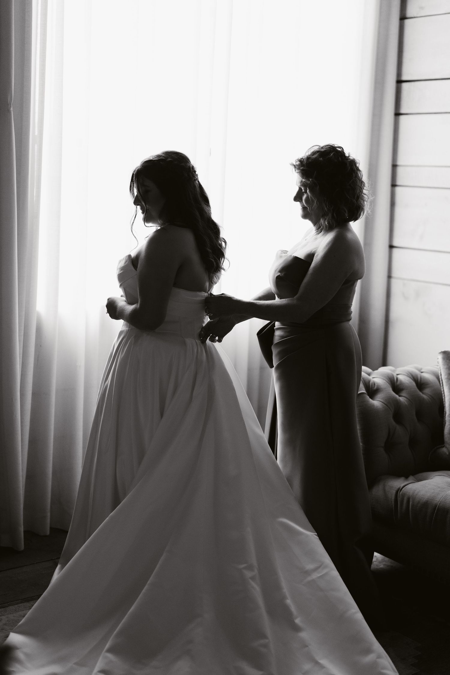 Black and white photo of bride in wedding gown being helped by bridesmaid near window with soft natural light.