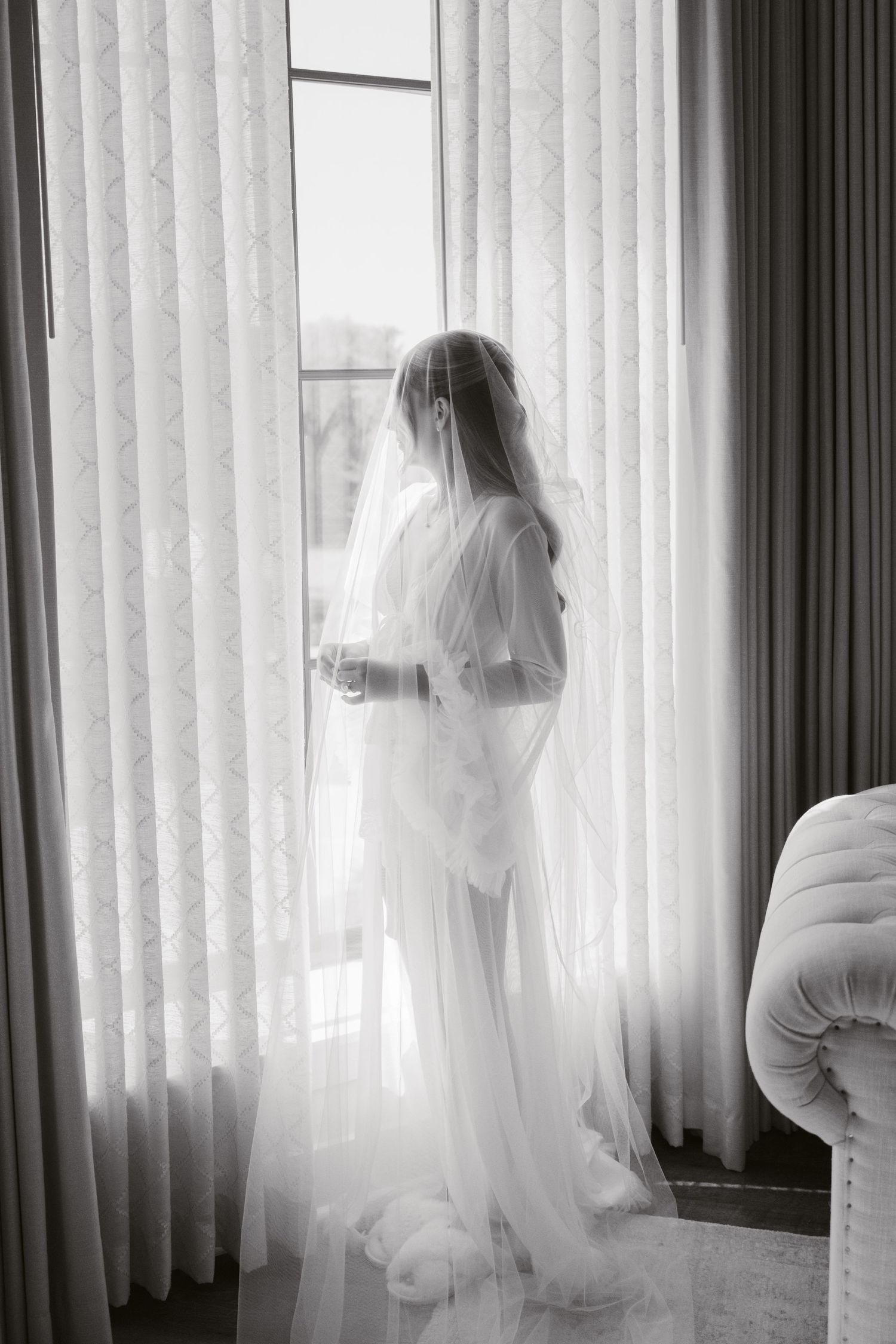 Black and white photo of a bride in a flowing white robe and veil standing by a large window on her wedding day.