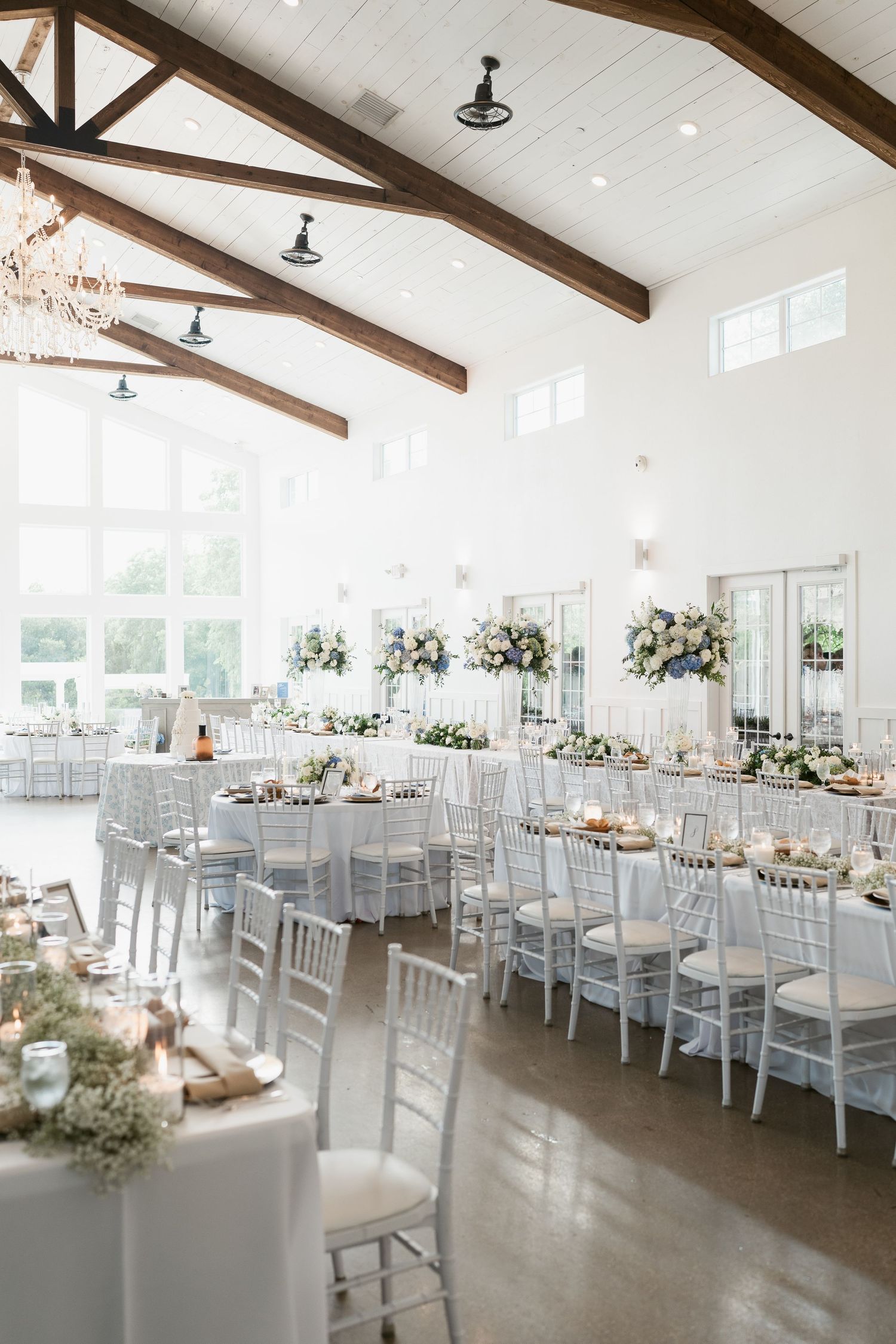 Elegant white wedding reception hall with vaulted wood beam ceilings, chiavari chairs, long tables, and floral centerpieces.