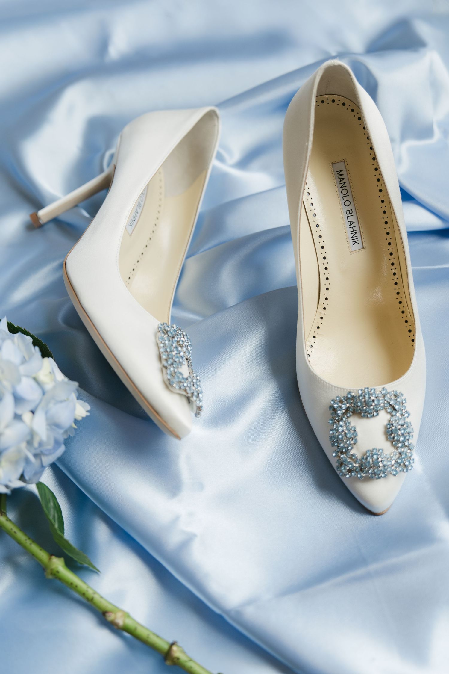 Elegant white Manolo Blahnik bridal heels with crystal embellishments on blue satin fabric with white flowers.