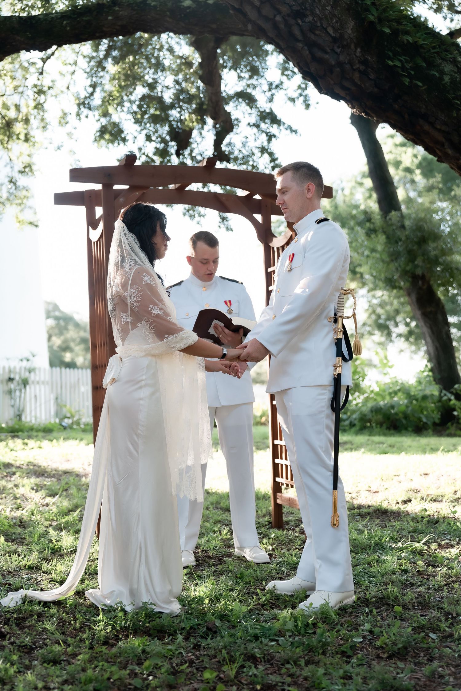 Military groom and bride exchange vows outdoors under wooden arch, officiant in white uniform, lush green trees backdrop.