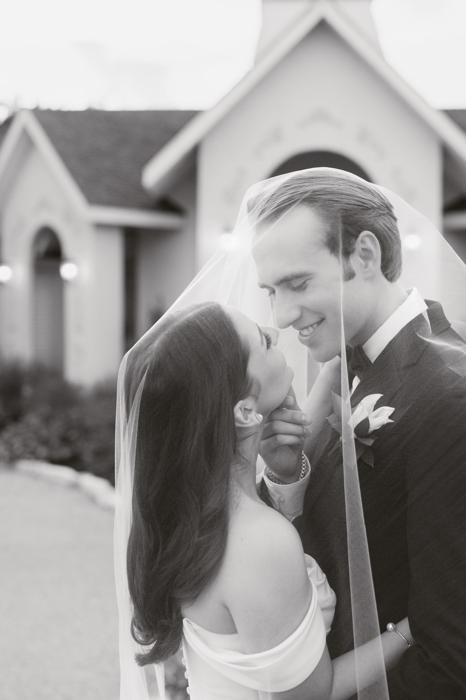 Black and white photo of a bride and groom sharing a romantic moment under a veil outside a chapel.