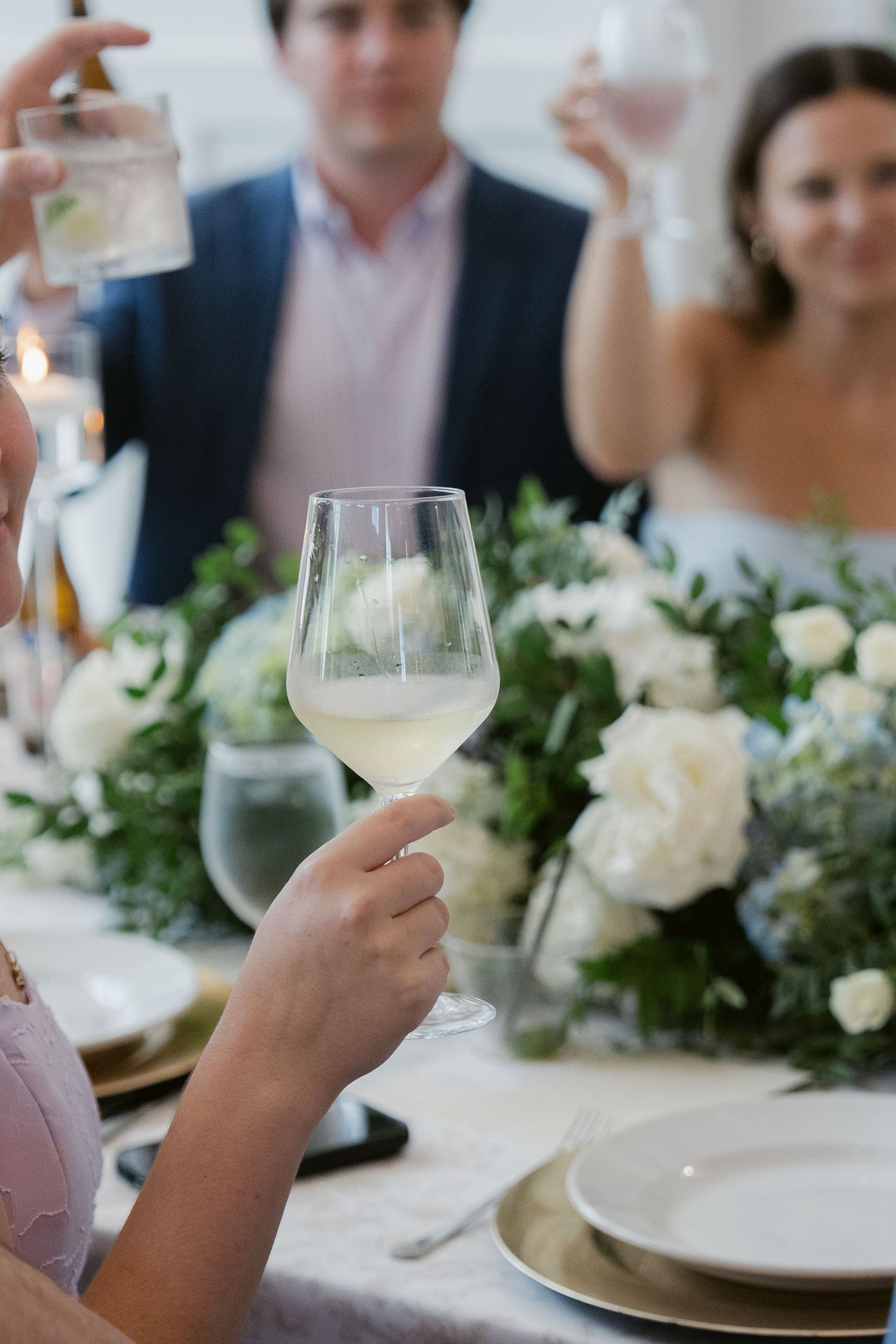 Guests raising glasses of white wine in a toast at an elegant wedding reception with white floral centerpieces.