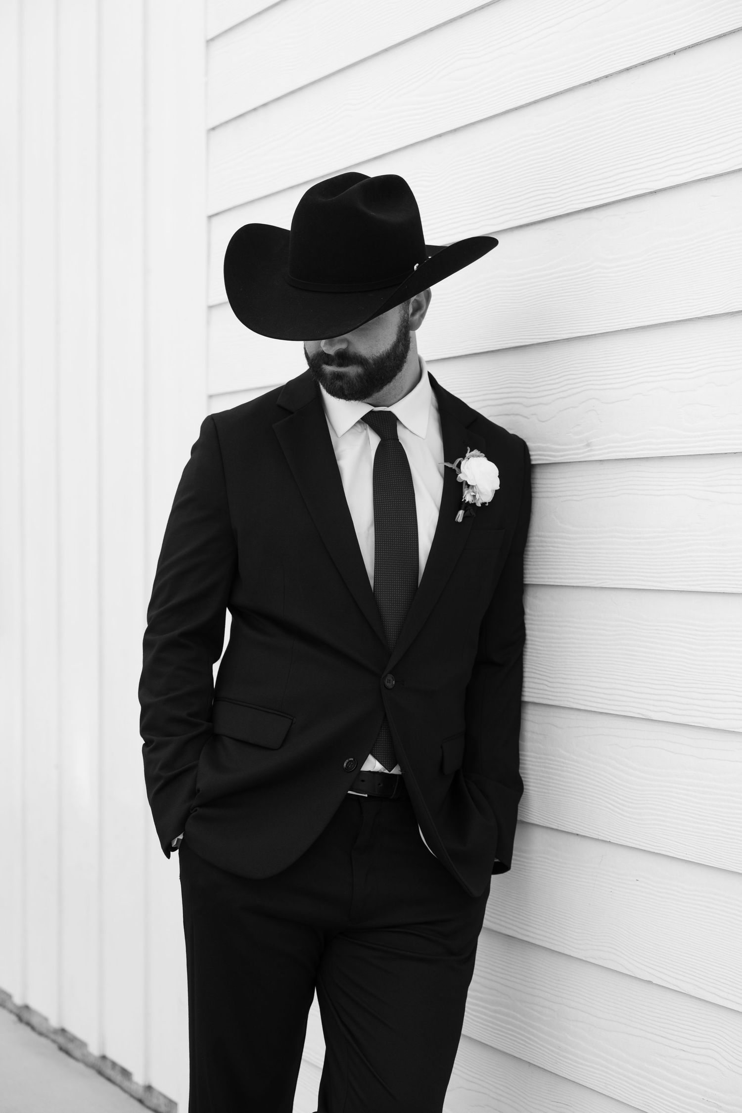 Bearded groom in black suit and cowboy hat with boutonniere leans against white wall.