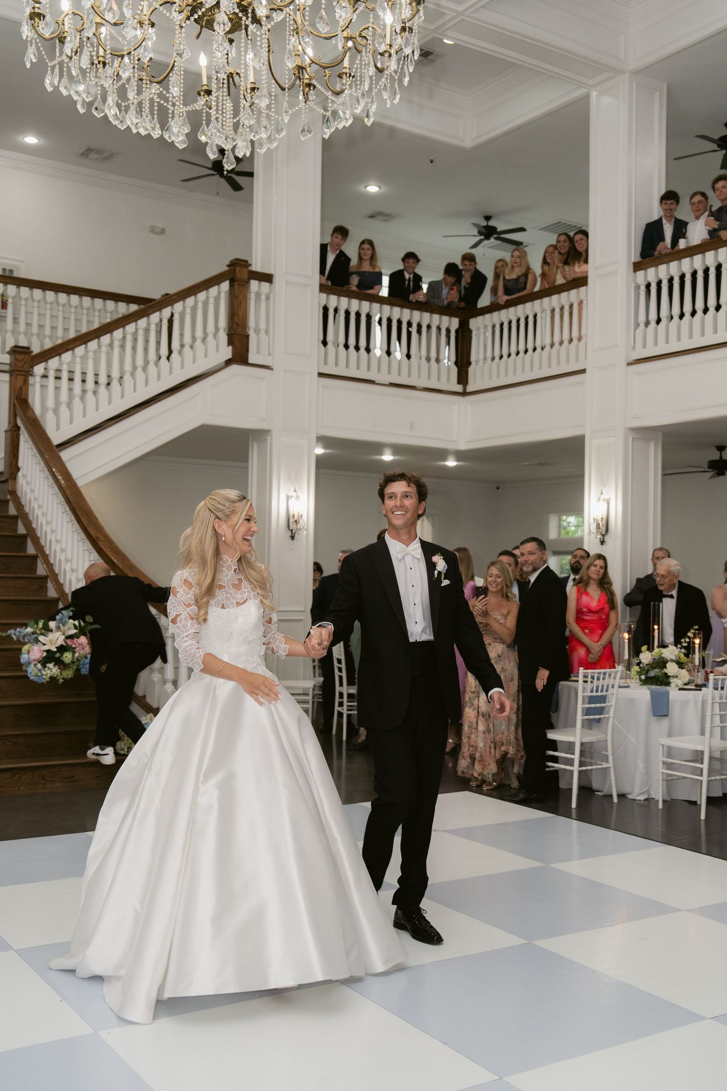 Bride and groom hold hands entering their elegant wedding reception in a grand ballroom with chandelier and staircase.