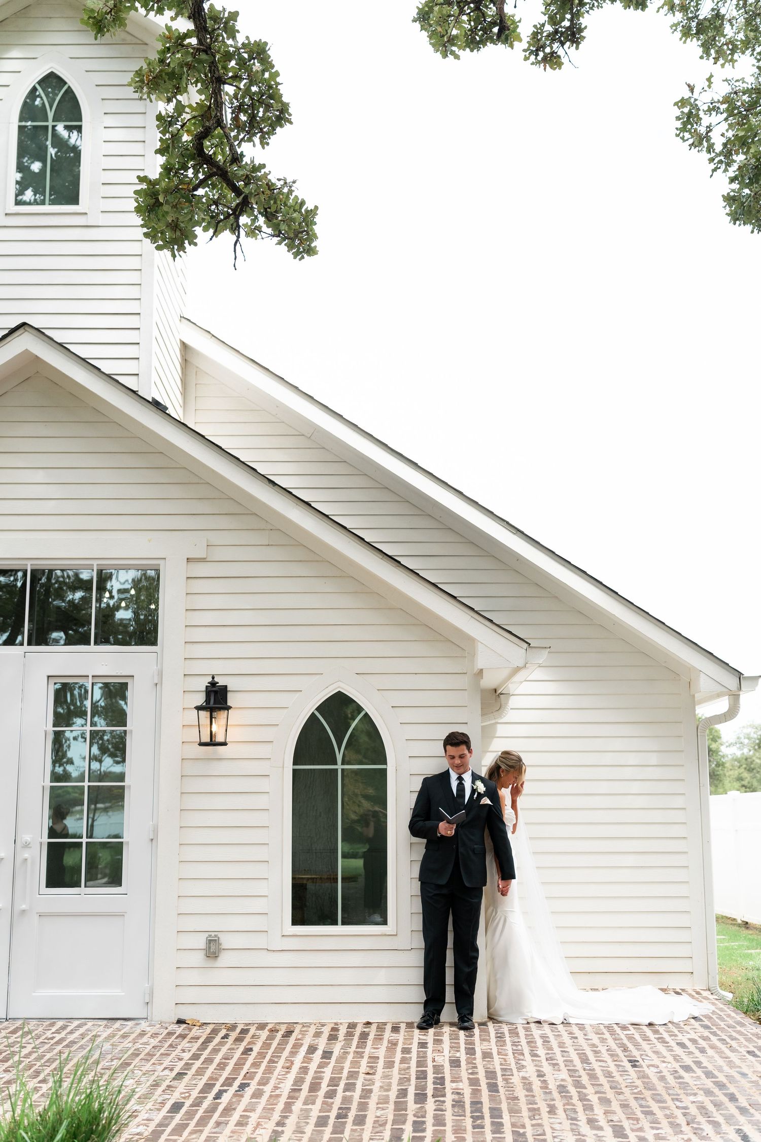 Bride and groom posing outside a white chapel with Gothic windows on a brick path on their wedding day