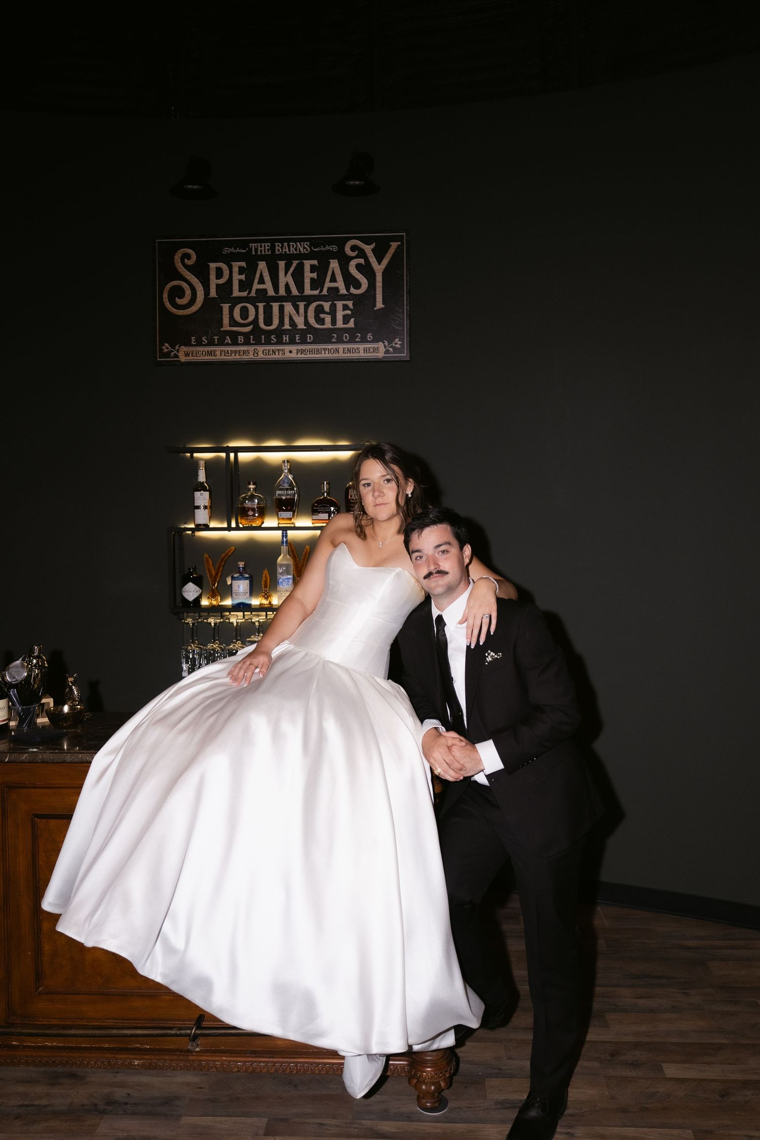 Bride in white gown sits on bar at Speakeasy Lounge while groom in black tuxedo poses beside her.