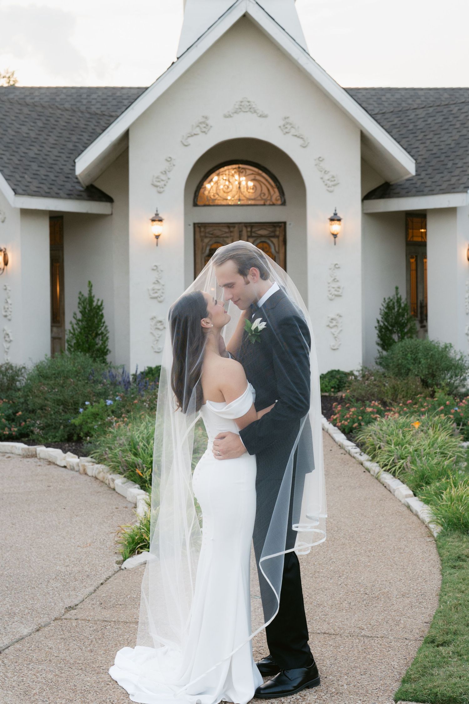 Bride and groom share a romantic kiss under a flowing veil outside an elegant white chapel at dusk.
