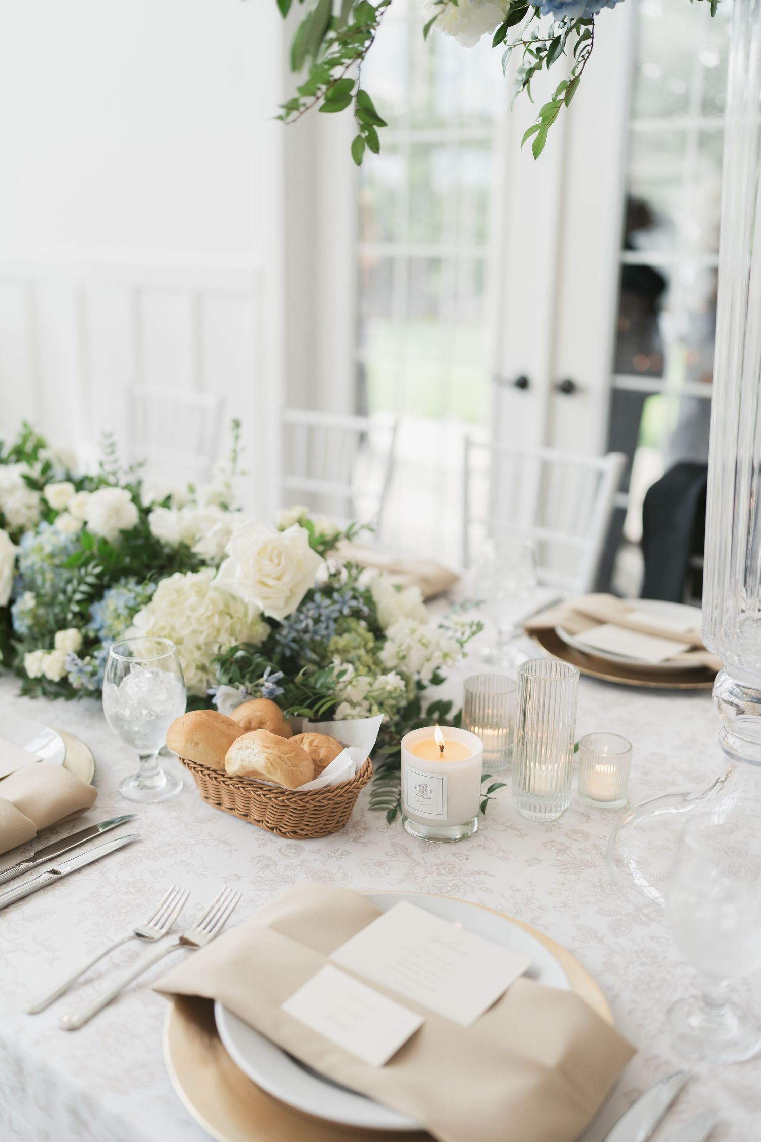 Elegant wedding reception table with white floral centerpiece, candles, bread basket, and neutral linen napkins on lace tablecloth.