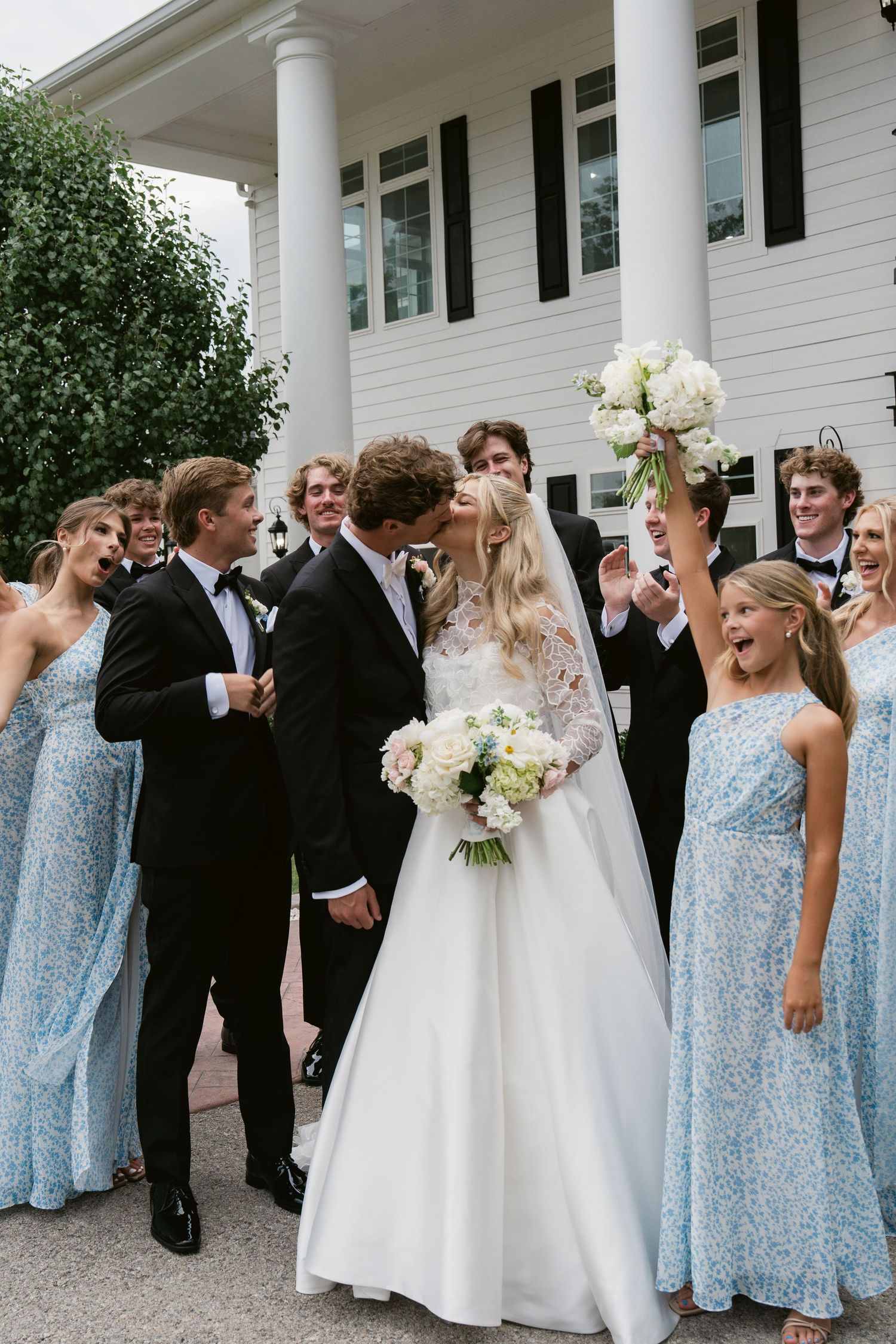 Wedding party celebrates outside white venue, bride and groom kiss as bridesmaids in blue dresses and groomsmen in tuxedos cheer.