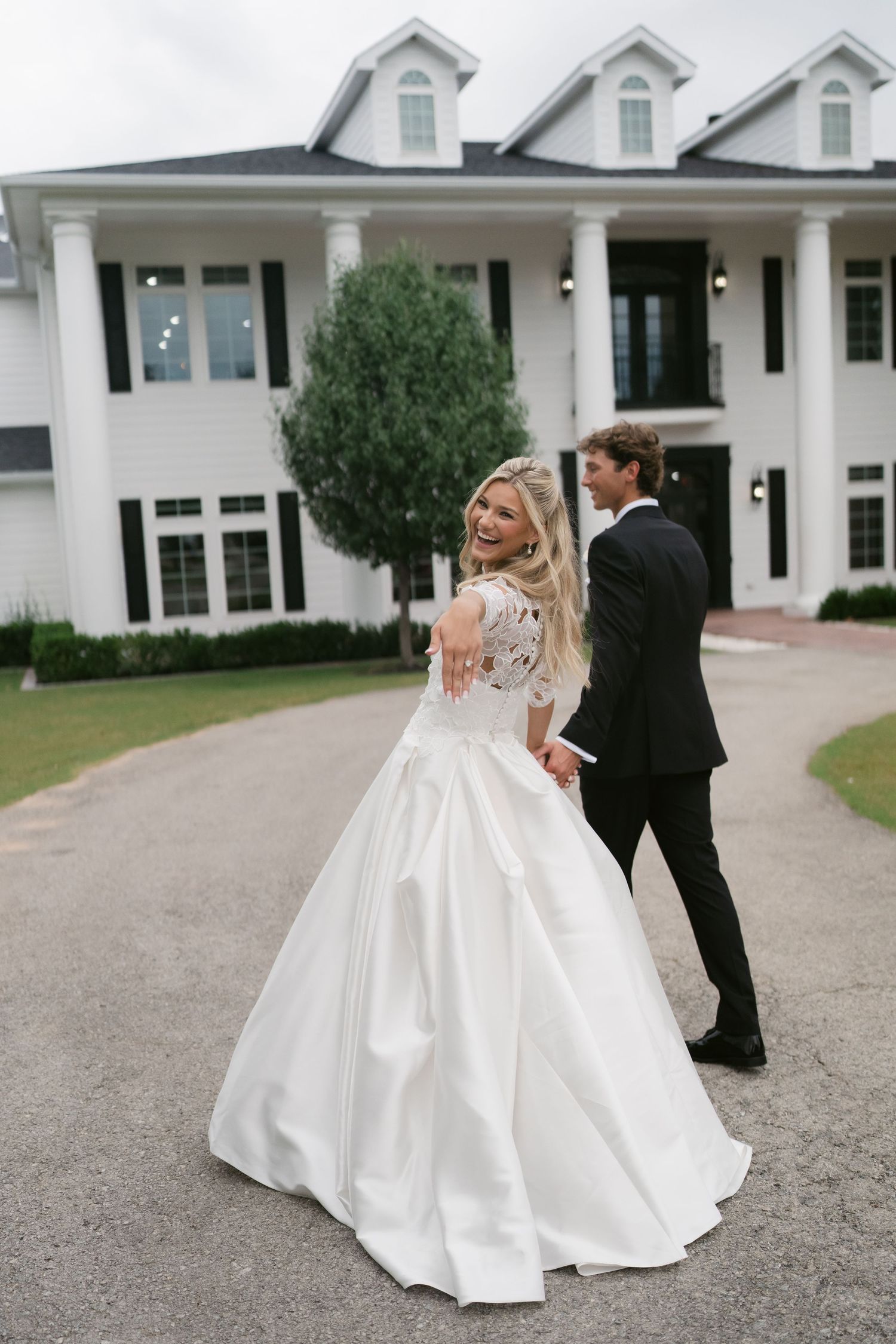 A bride in a white ball gown laughs over her shoulder while holding hands with a groom in a black tuxedo outside a white mansion.