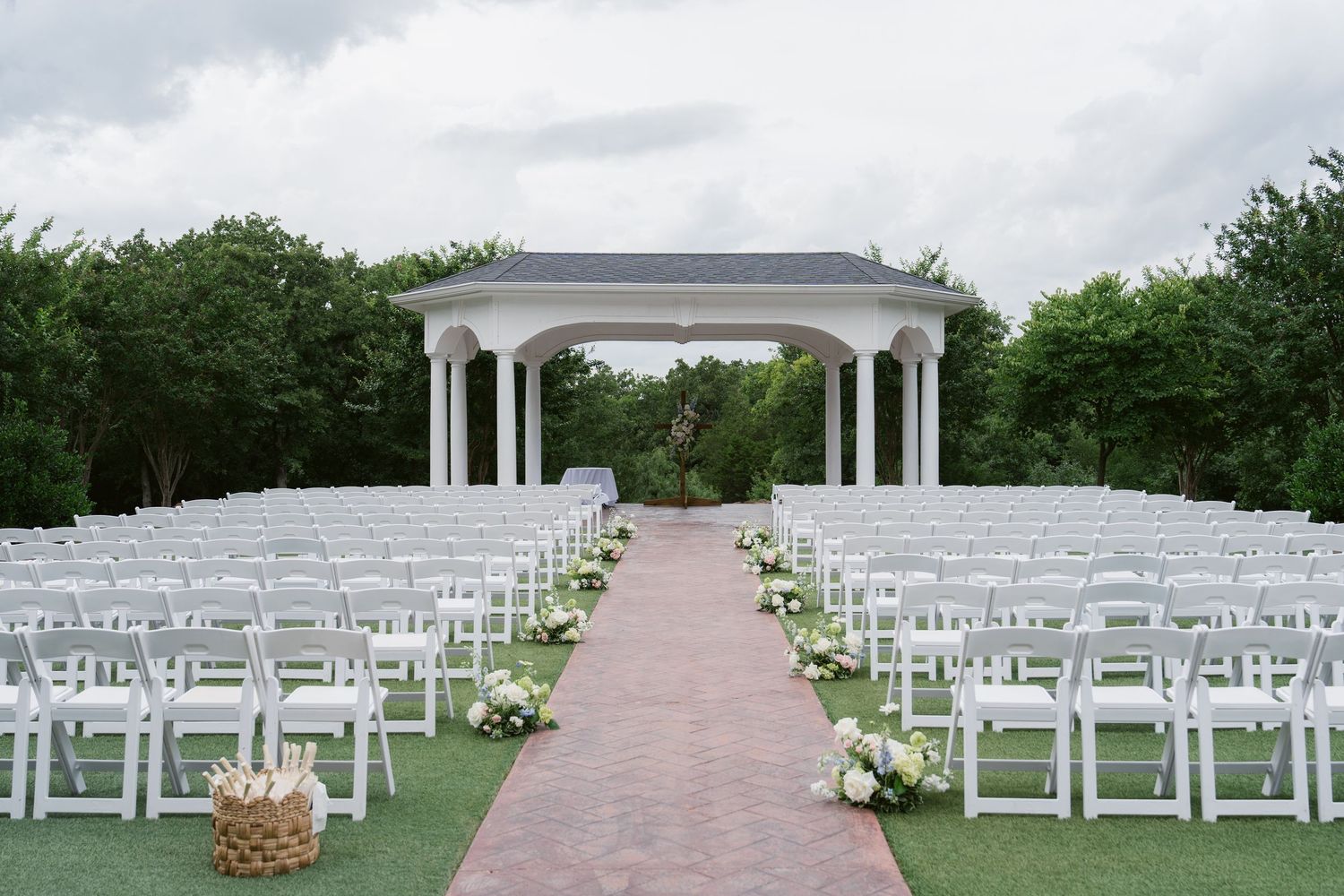 Elegant outdoor wedding ceremony setup with white chairs, floral aisle decorations, and a classic white gazebo surrounded by lush greenery.