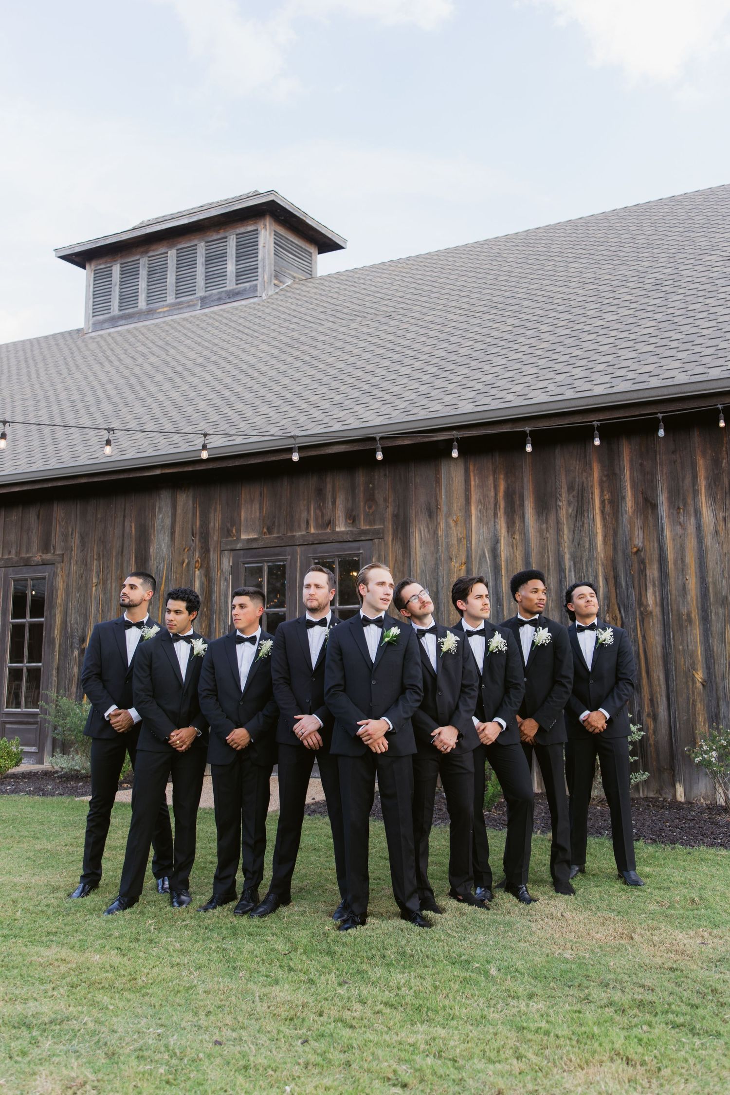 Groomsmen in black tuxedos and bow ties pose together on a lawn in front of a rustic wooden barn venue.