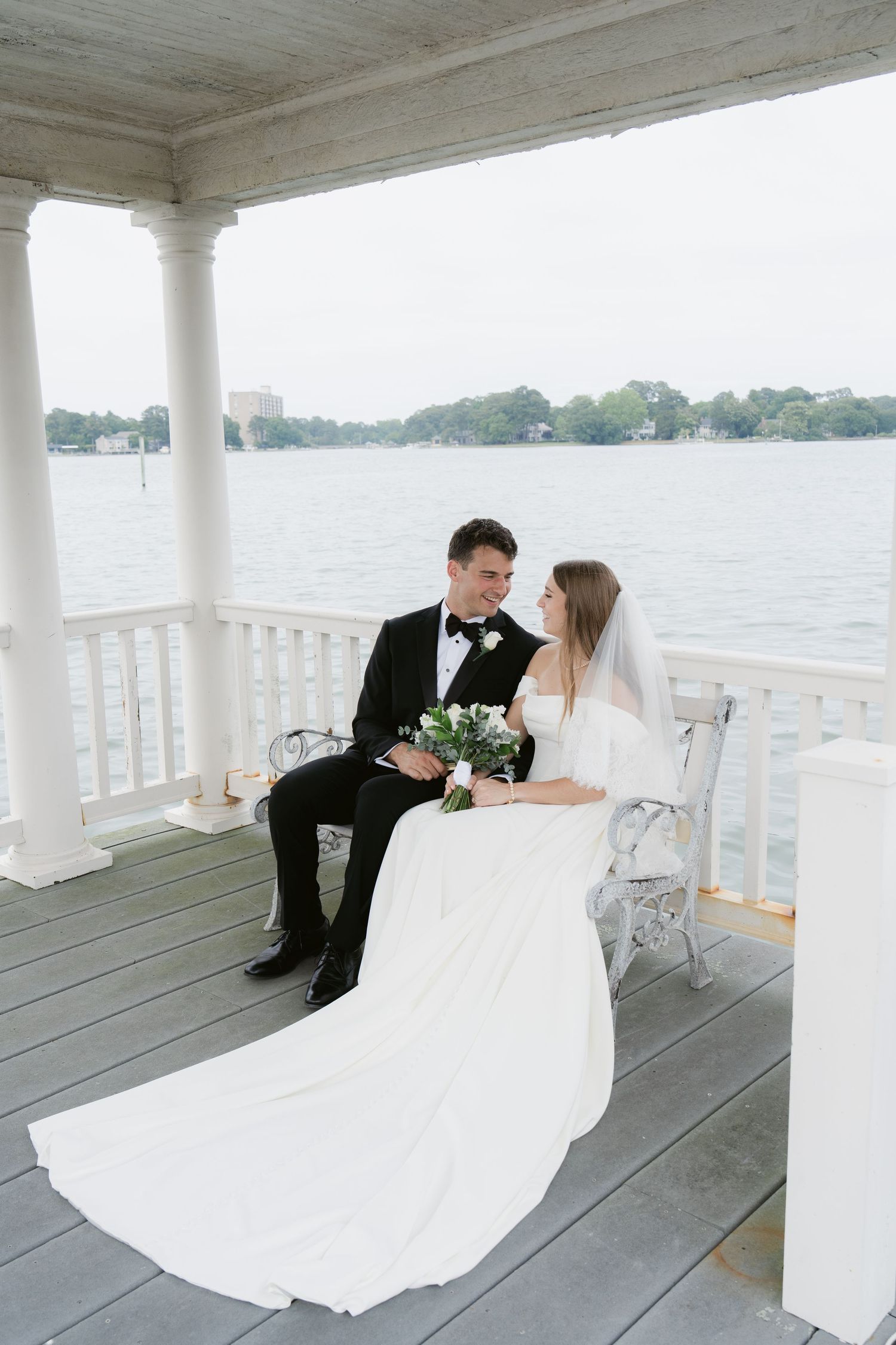 Bride and groom sitting together on a waterfront gazebo, bride in white gown holding bouquet, groom in black tuxedo in Norfolk, Virginia