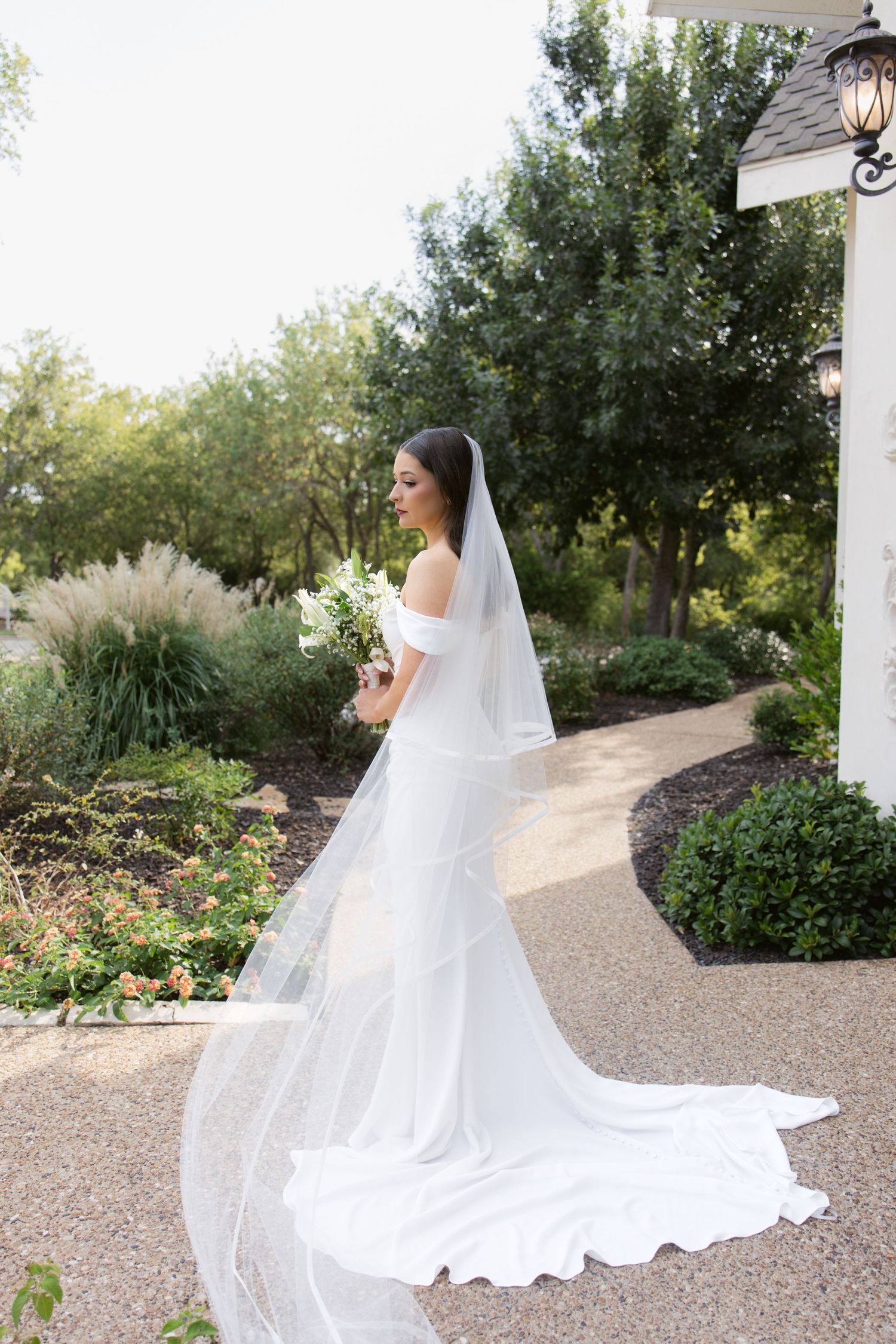 Bride in white off-shoulder gown with long cathedral veil holding bouquet in lush garden setting.