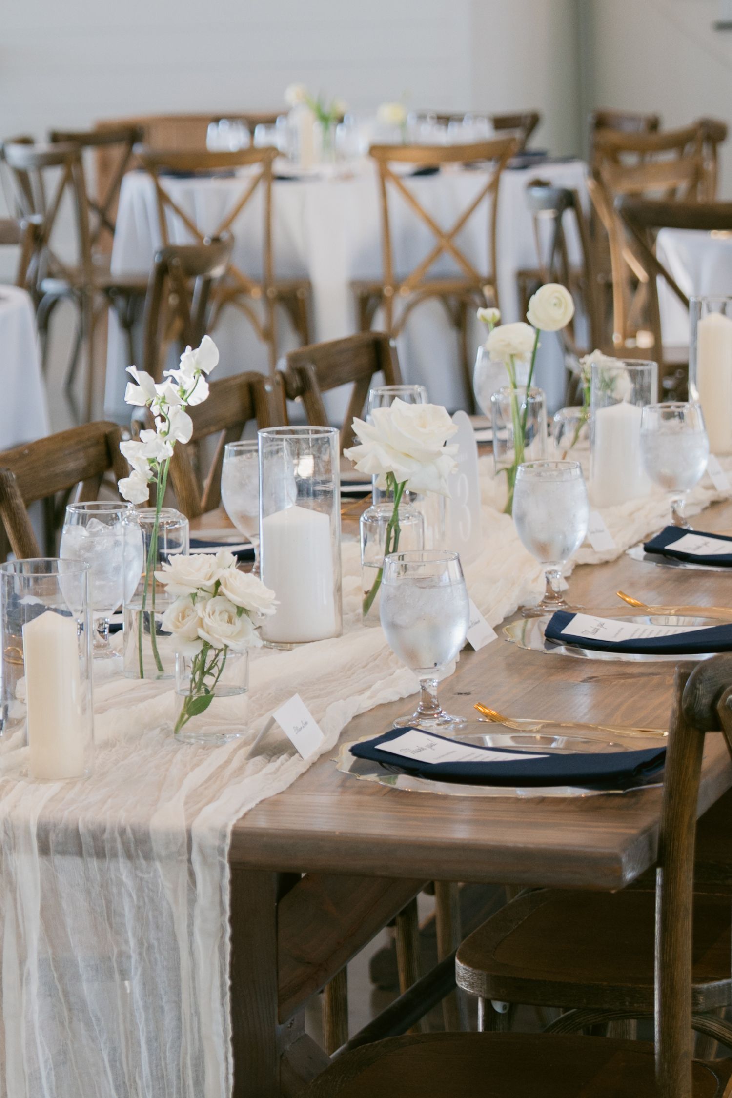 Elegant wedding reception table with white floral centerpieces, candles, navy napkins, and a neutral linen runner on wood tables.