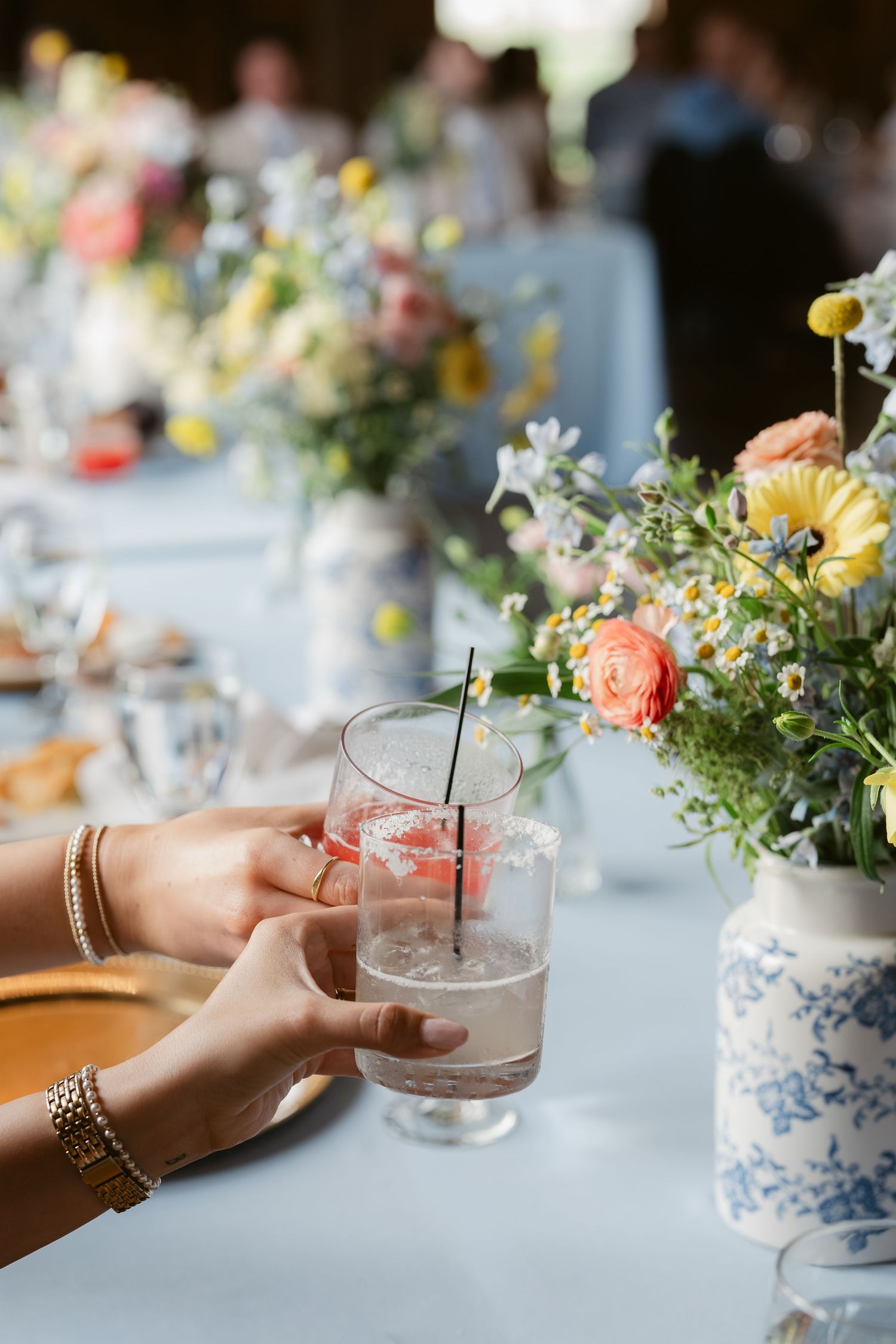 Two hands clinking cocktail glasses at an elegant wedding reception table adorned with colorful spring floral arrangements.