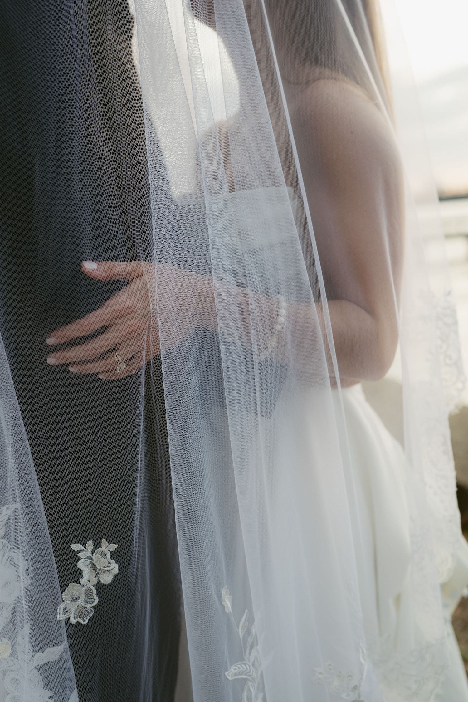 Bride in white gown and floral veil embraces groom in dark suit, pearl bracelet and wedding ring visible.