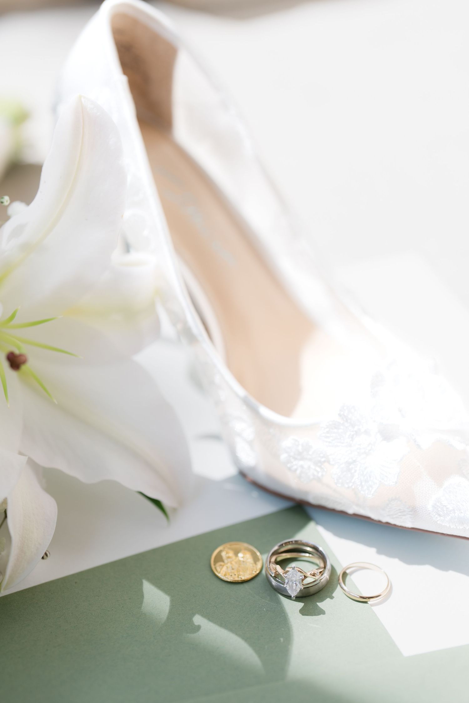White floral bridal heels displayed beside wedding rings, engagement ring, and gold coin on a sage green surface.