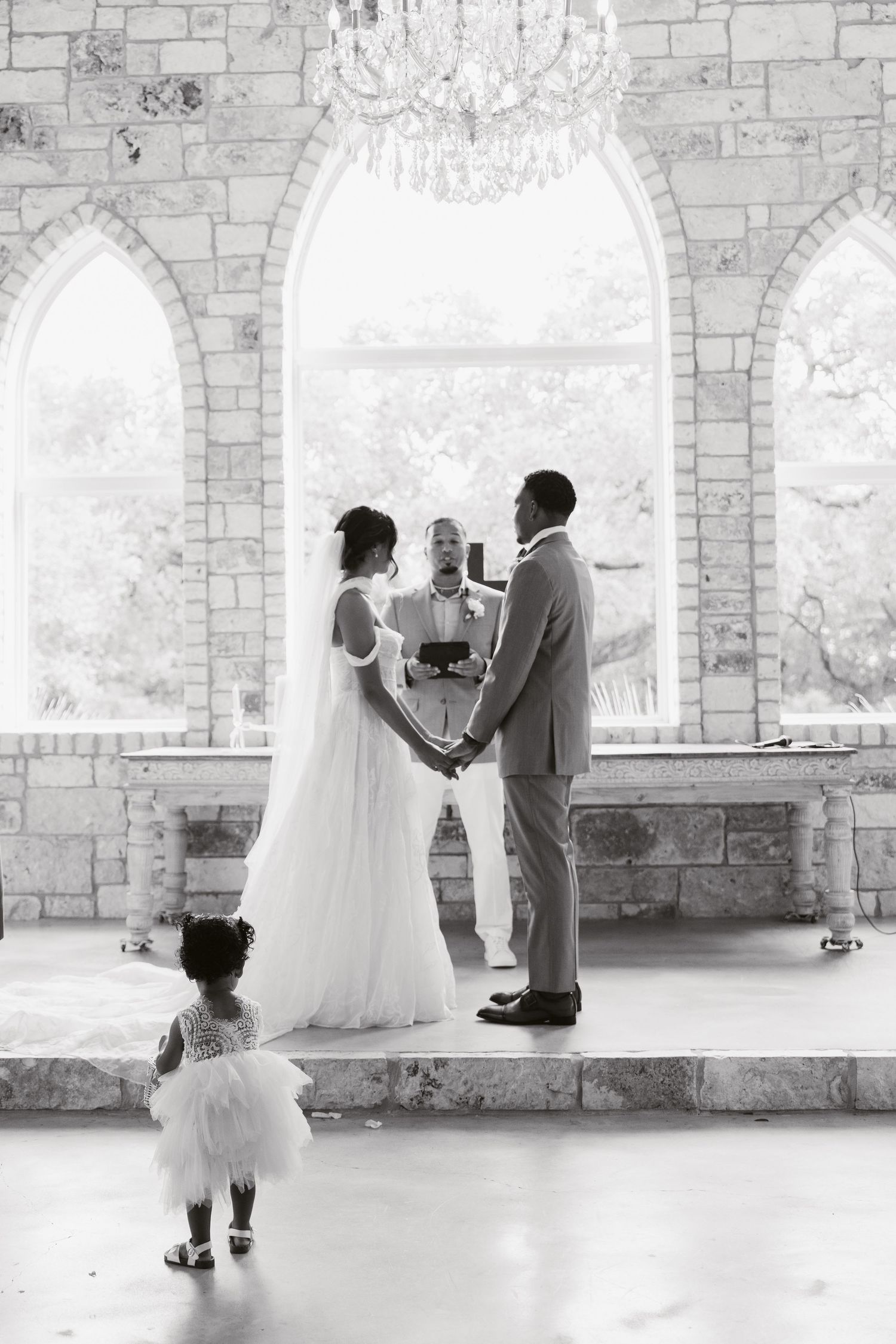 Black and white wedding ceremony photo with couple holding hands before officiant in elegant stone chapel with chandelier.