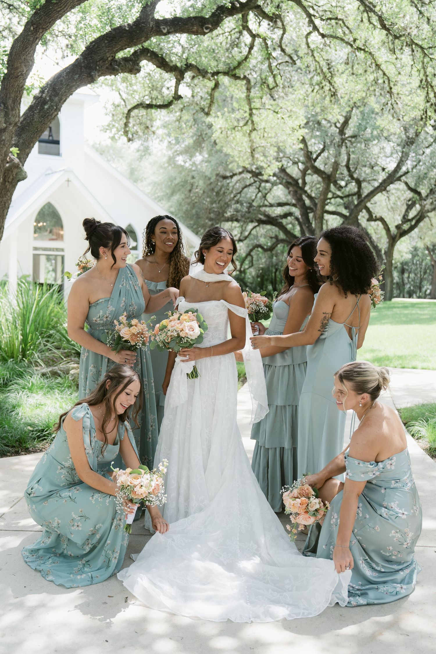 Bride in white gown surrounded by bridesmaids in sage green dresses holding peach bouquets outdoors near white chapel.