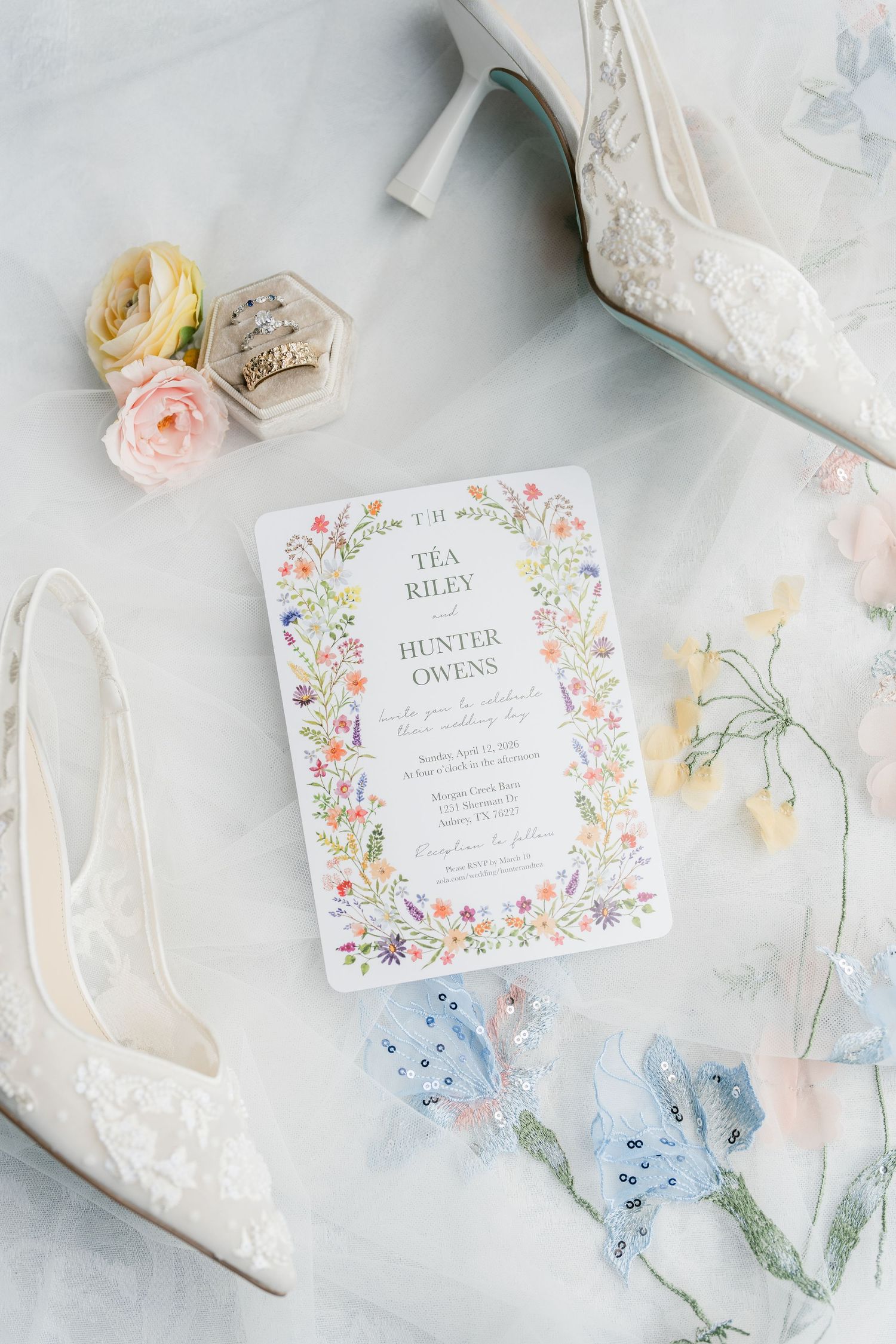 Elegant wedding invitation with colorful floral border surrounded by bridal heels, pink roses, and delicate accessories.