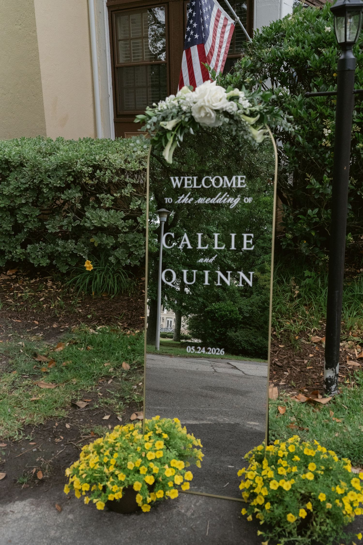 Mirror wedding welcome sign for Callie and Quinn's May 24 2026 wedding, adorned with white floral arrangement and yellow flowers.