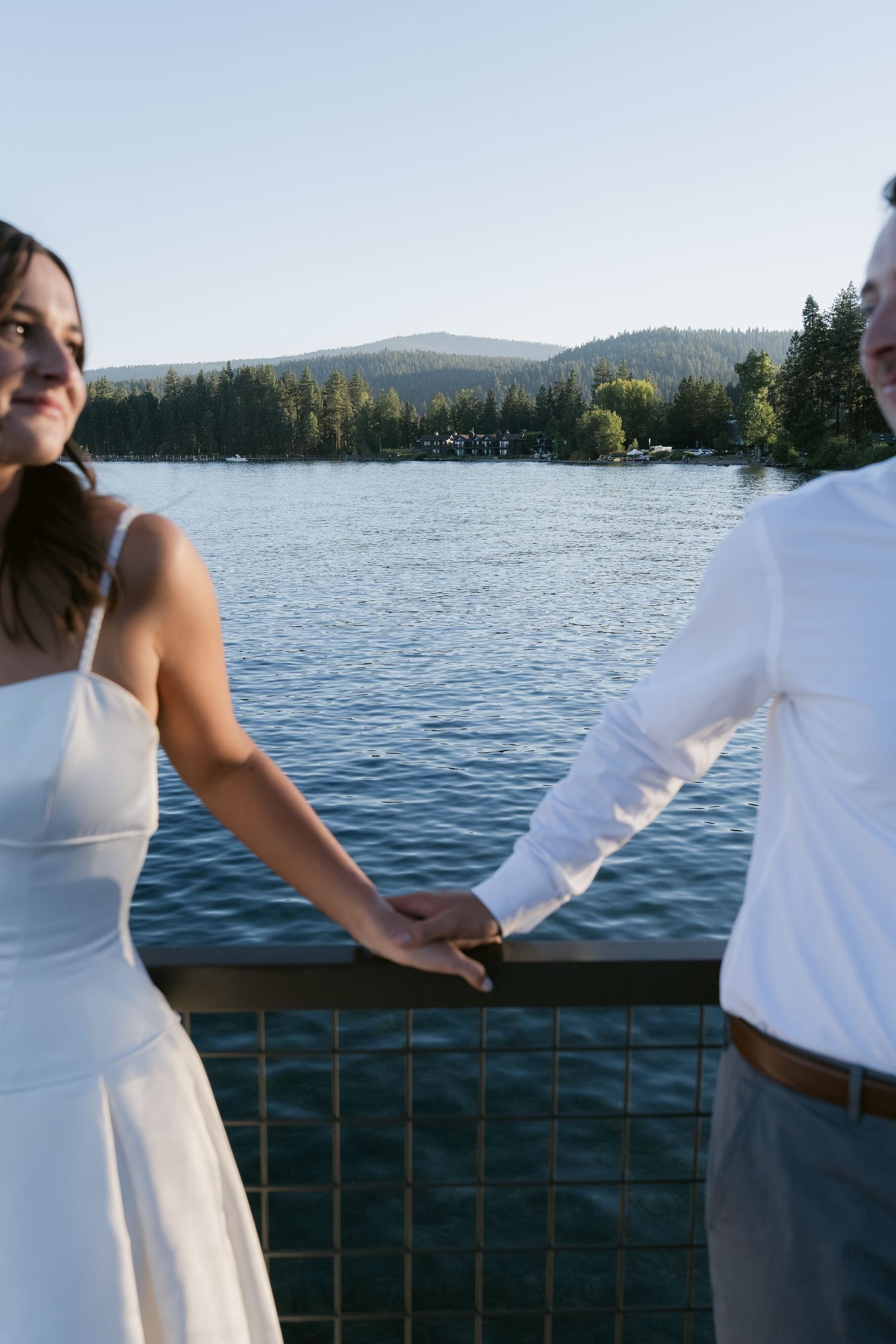 Couple holding hands on a lakeside deck with forested mountains in the background on a sunny day.