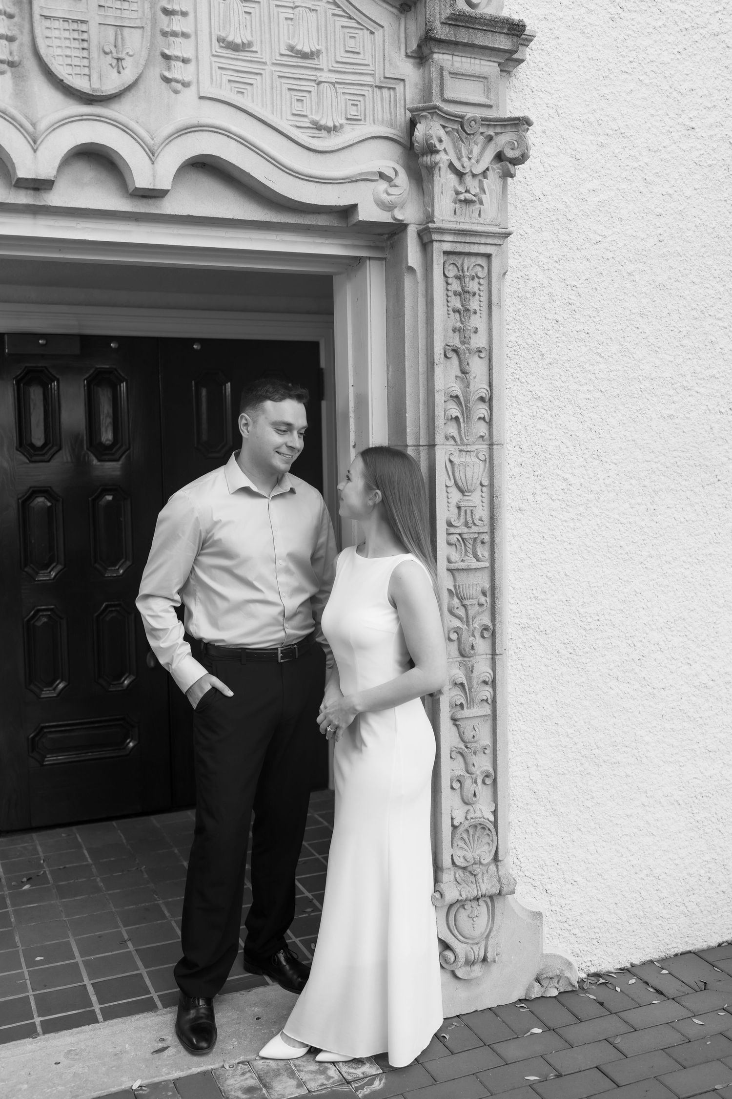 Black and white photo of a couple in formal attire standing by an ornate church doorway, sharing a romantic moment.