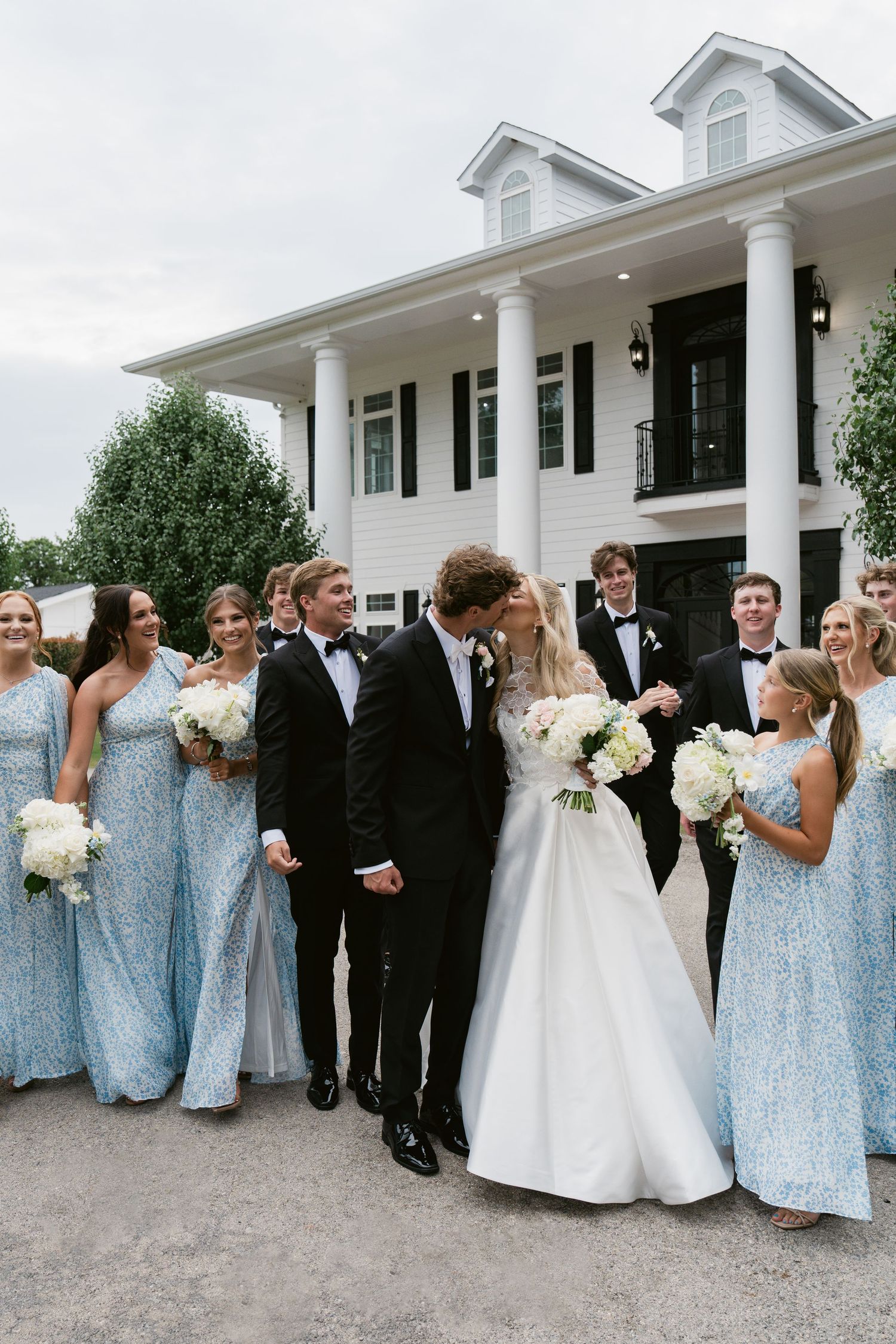 Wedding party poses outside white colonial venue, bride in white gown with bridesmaids in light blue dresses and groomsmen in tuxedos.