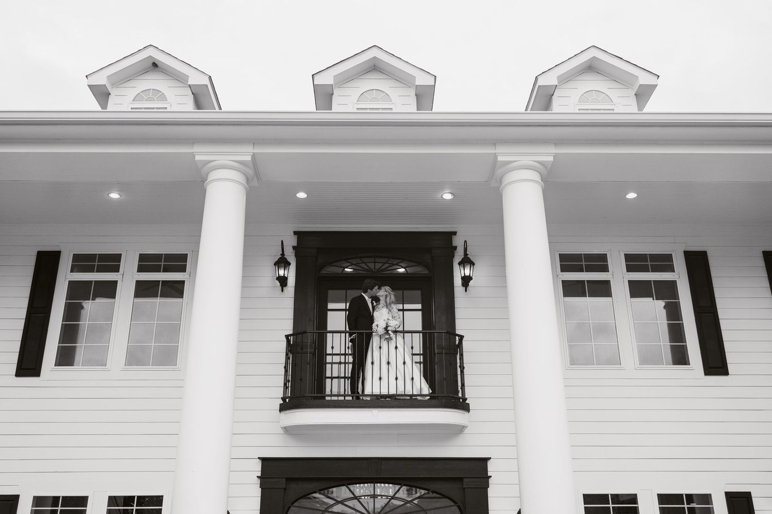 Black and white photo of a couple kissing on a balcony of an elegant white colonial mansion with tall columns.