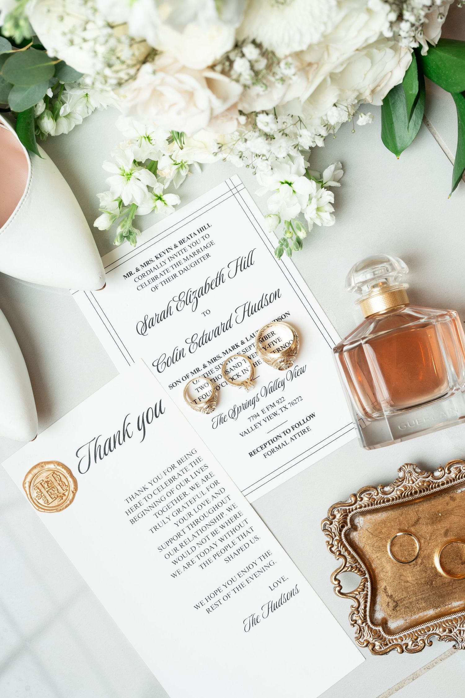 Elegant wedding flat lay with floral invitation suite, gold rings, perfume bottle, and white roses bouquet.