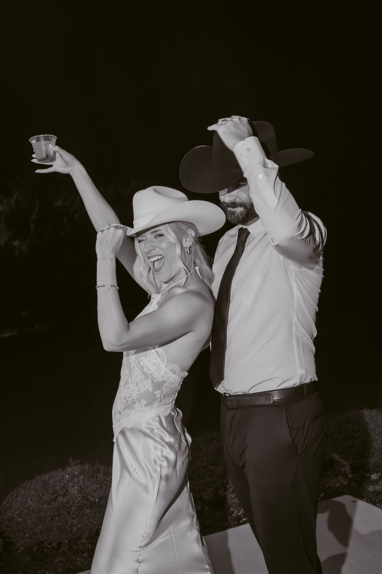 Black and white photo of a smiling bride and groom in cowboy hats dancing joyfully at their wedding reception.