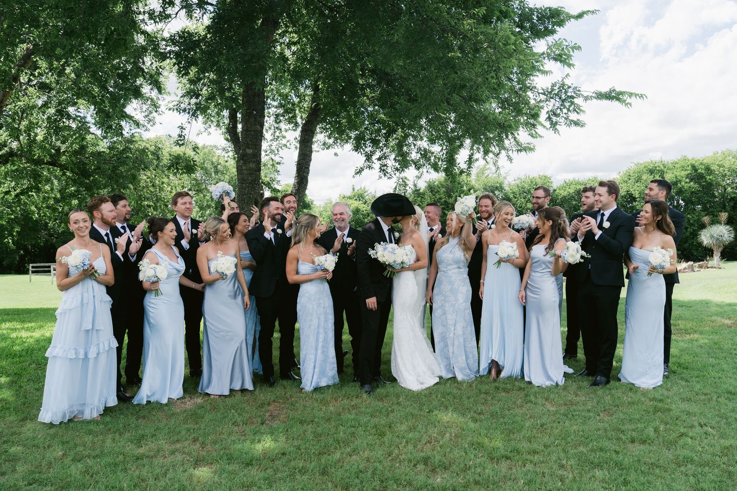 Large wedding party posing outdoors in garden, bridesmaids in light blue gowns, groomsmen in black tuxedos.