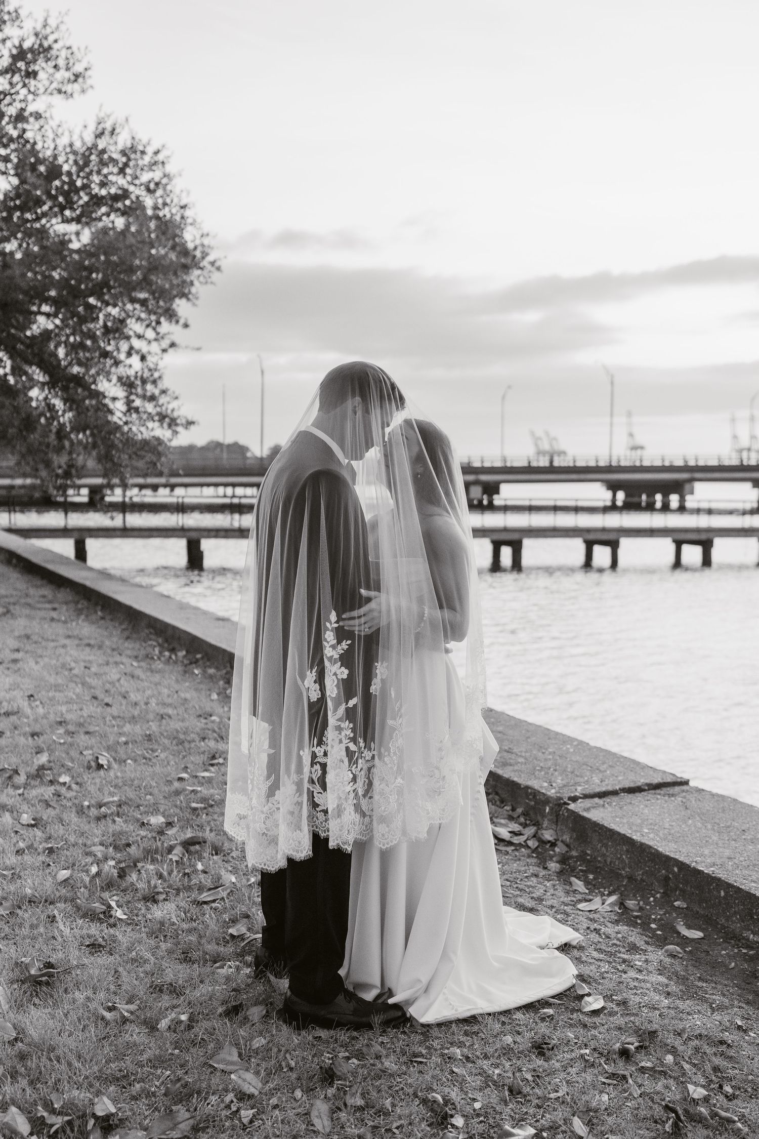 Black and white photo of bride and groom kissing under veil by waterfront pier in Norfolk, Virginia