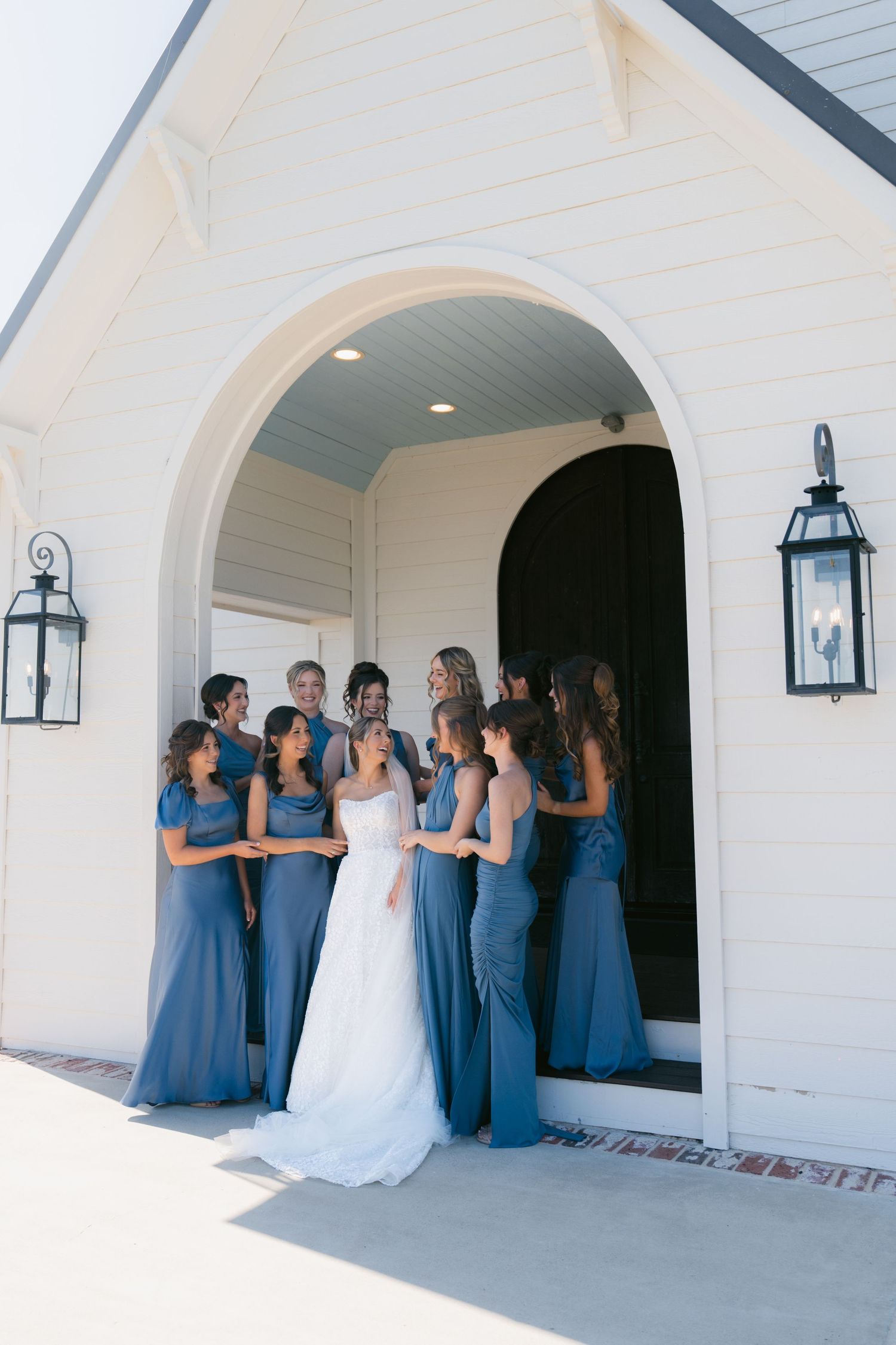 Bride in white gown surrounded by bridesmaids in dusty blue dresses at a white chapel entrance.