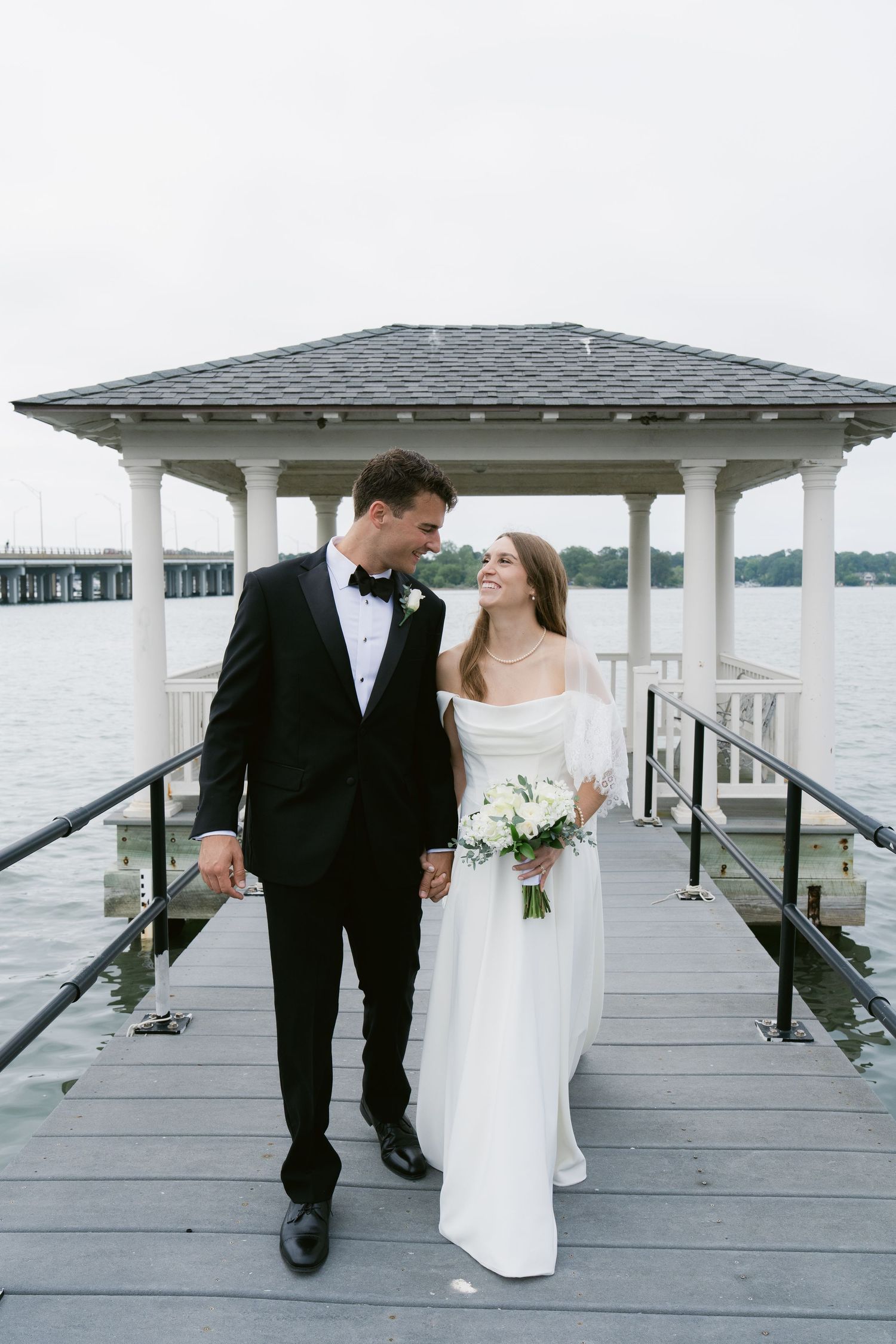 A bride and groom hold hands on a waterfront dock gazebo, she in a white gown holding flowers, he in a black tuxedo.