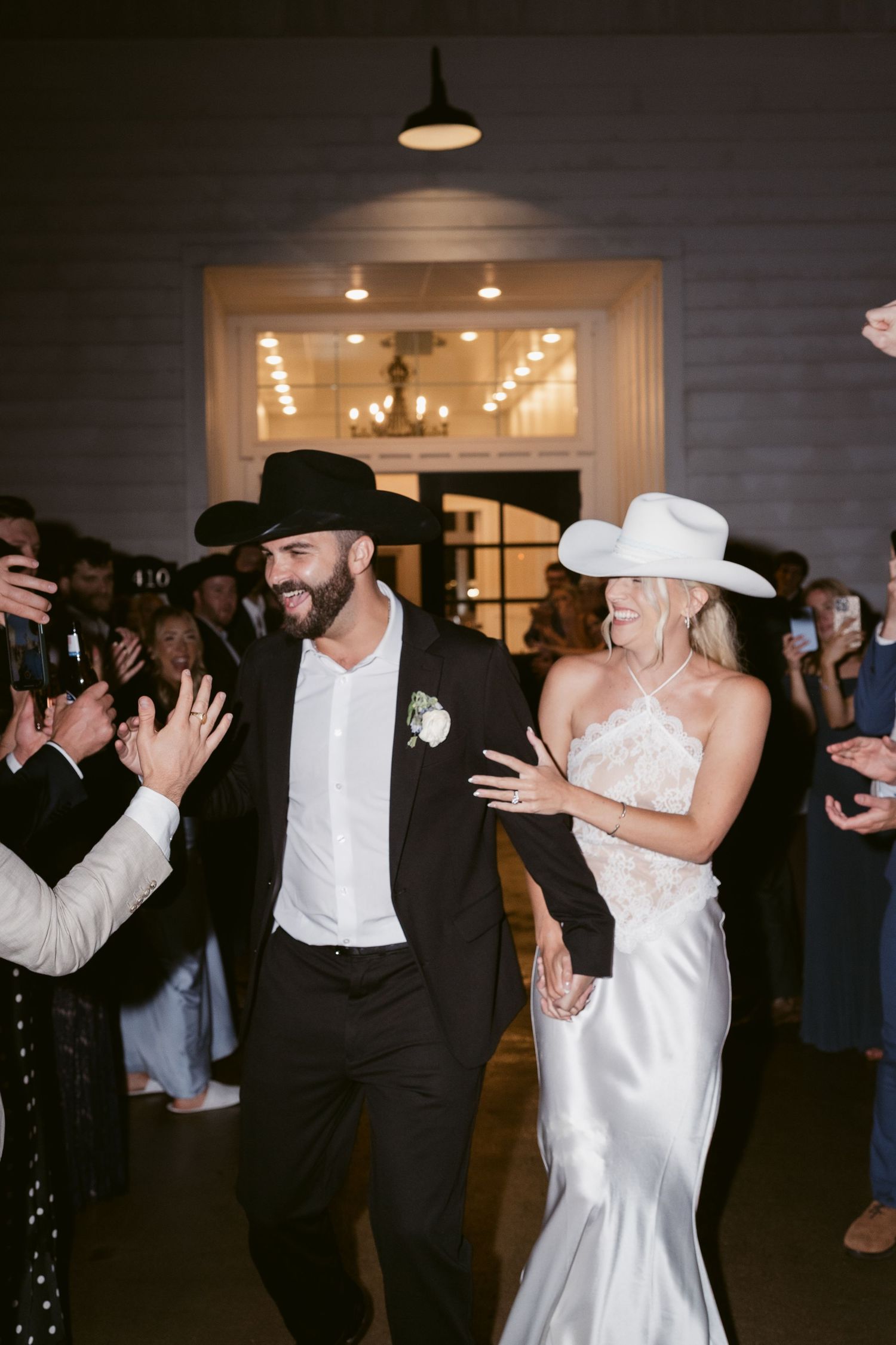 Smiling bride and groom in cowboy hats make grand entrance at wedding reception while guests cheer and applaud.