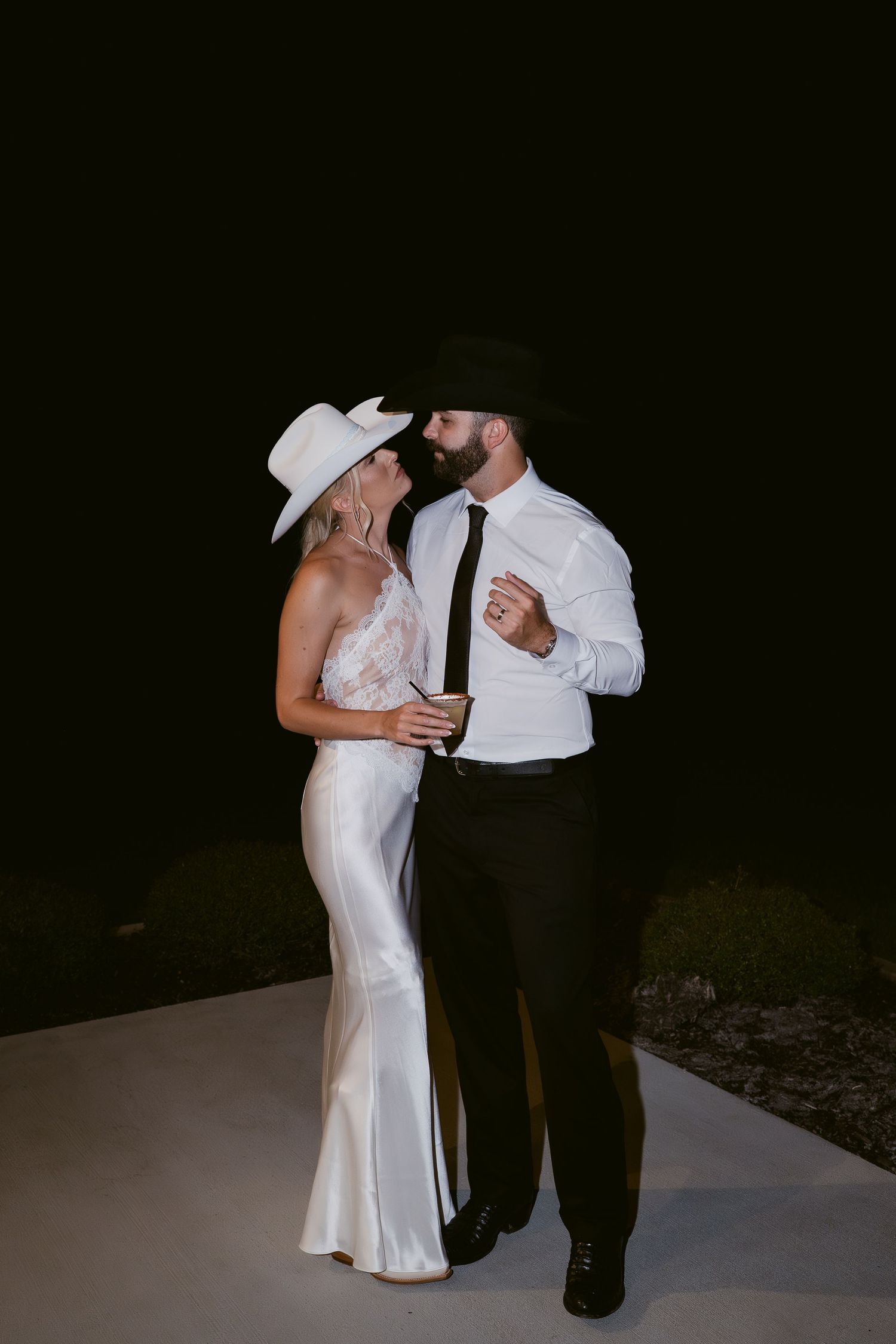 Couple in cowboy hats share romantic moment outdoors at night, bride in white satin gown.