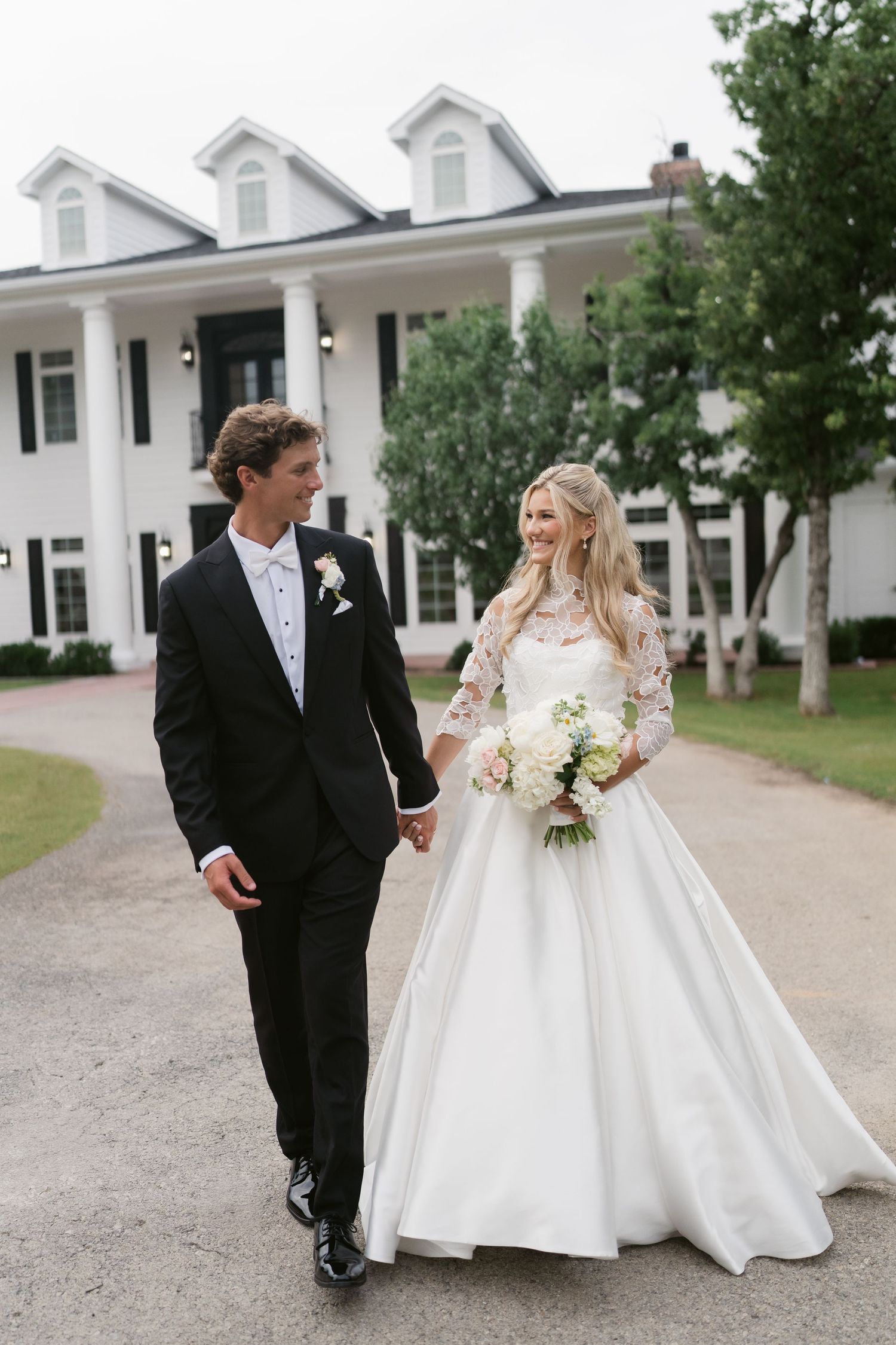 A bride and groom hold hands walking in front of a white colonial mansion on their wedding day.