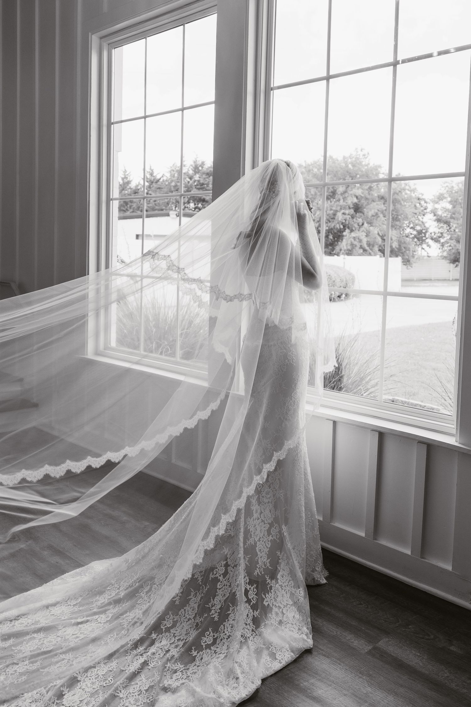 Black and white photo of a bride in a lace wedding gown with a flowing cathedral veil standing by large windows.