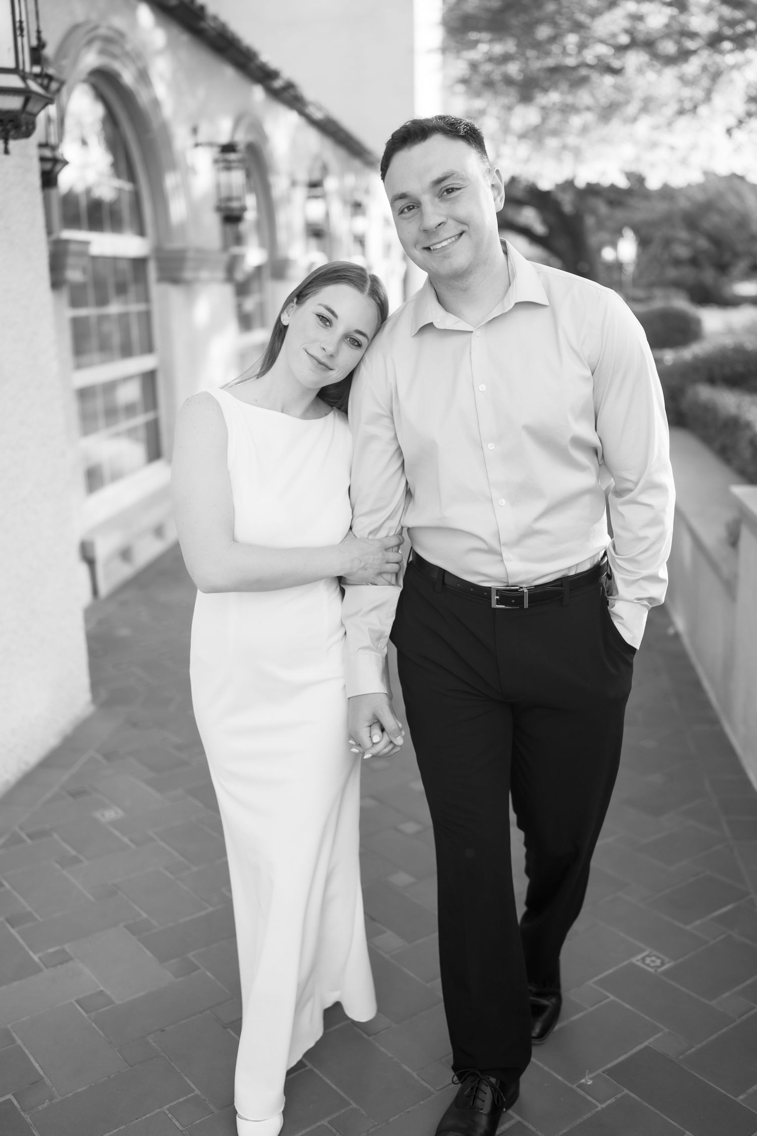 Black and white photo of a smiling couple in formal attire posing together on an elegant outdoor walkway.