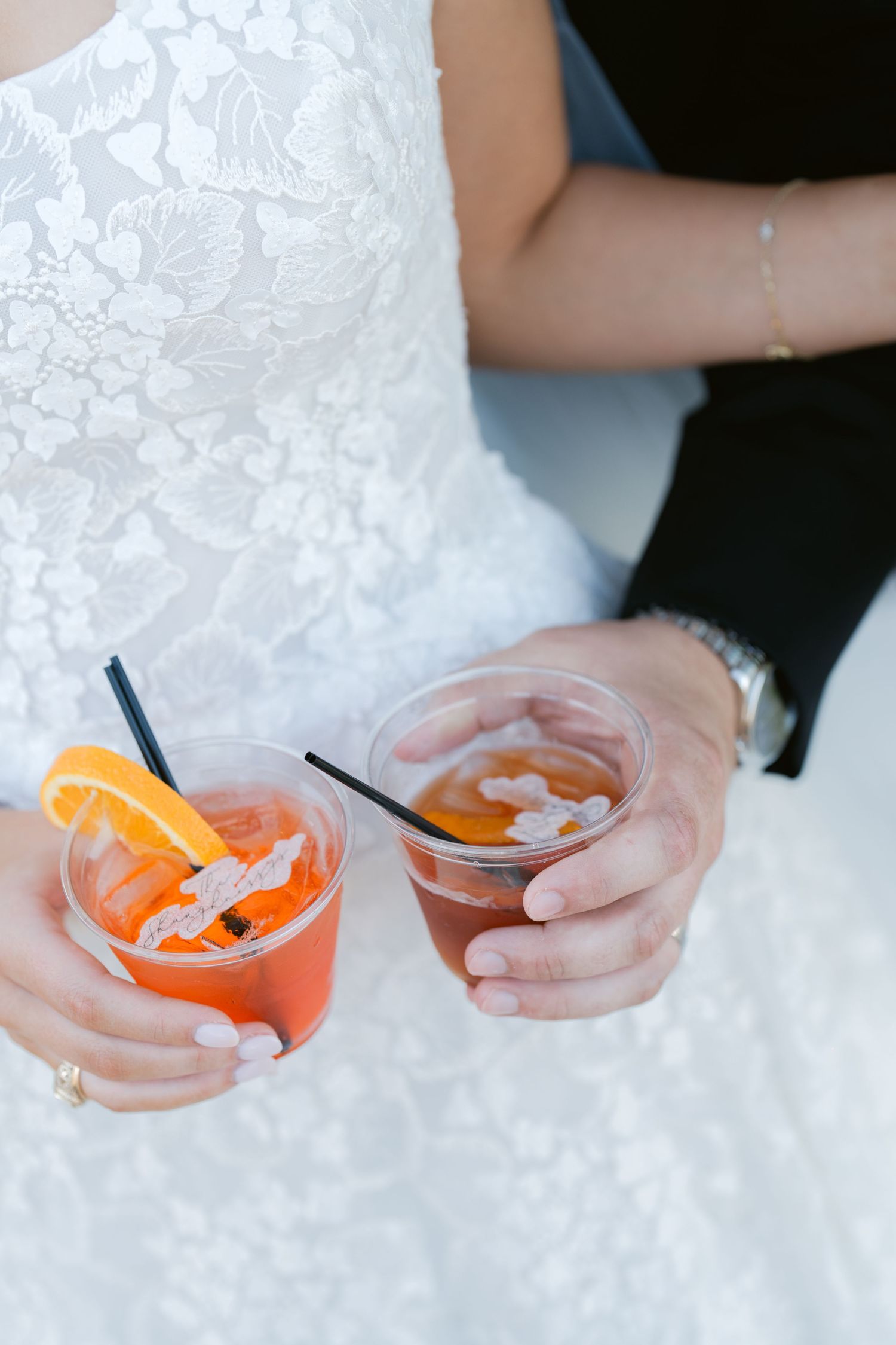 Hands holding orange cocktails garnished with citrus slices against white lace fabric at a wedding reception.