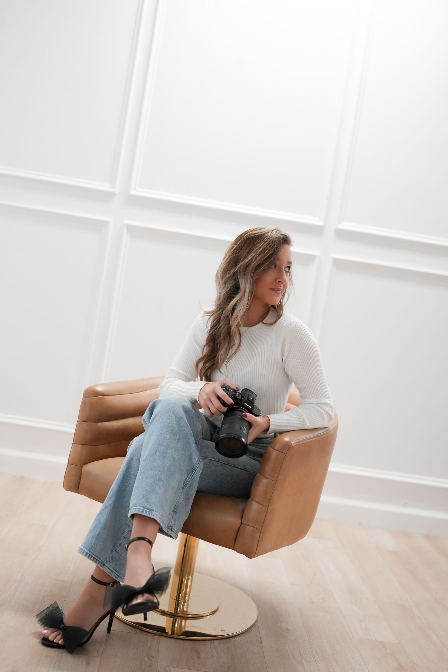 Photographer lounging on brown leather chair while holding camera in bright white studio space.