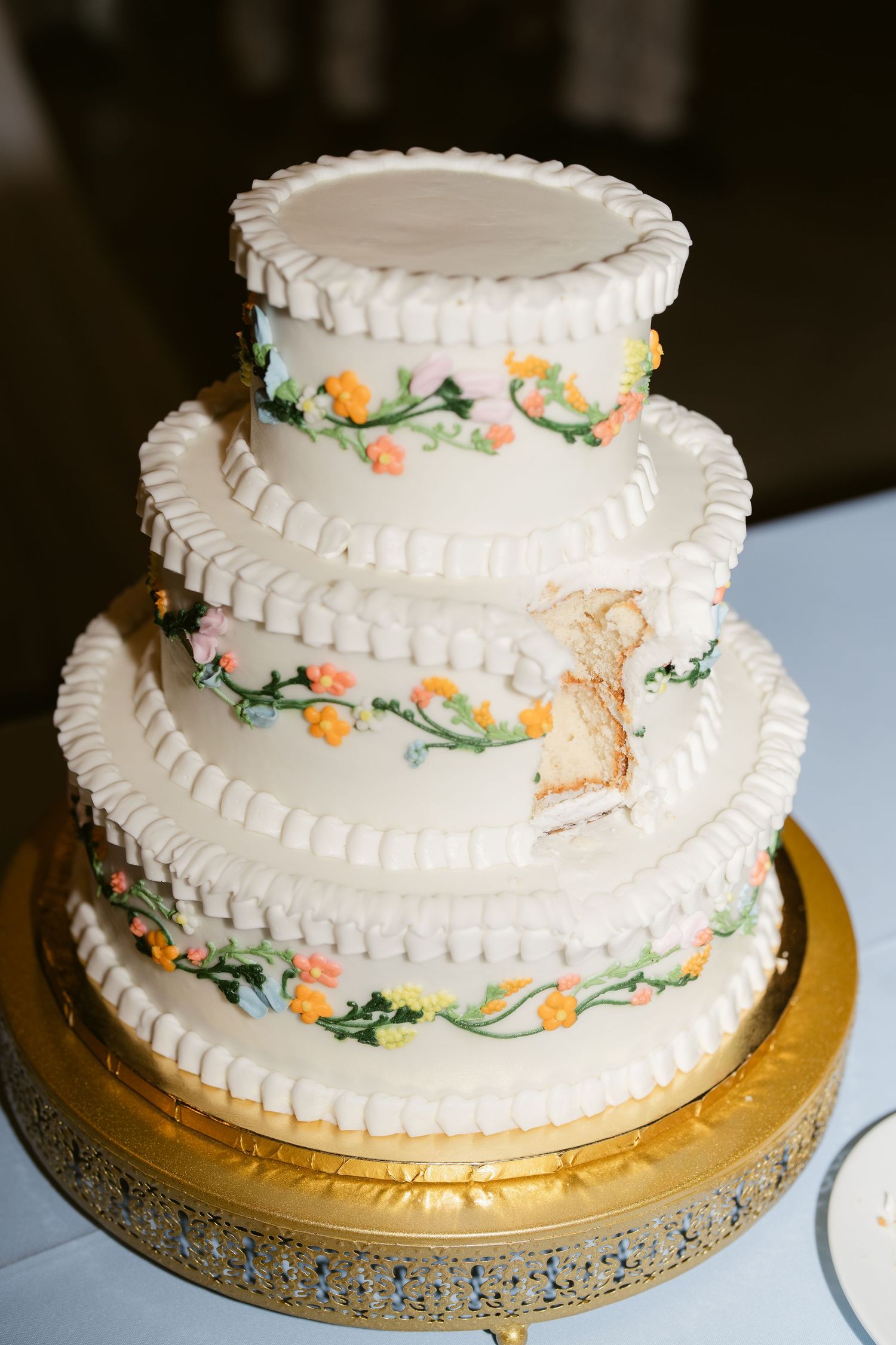 Three-tier white wedding cake with floral vine decorations and ruffled edges on a gold stand, with a slice removed.