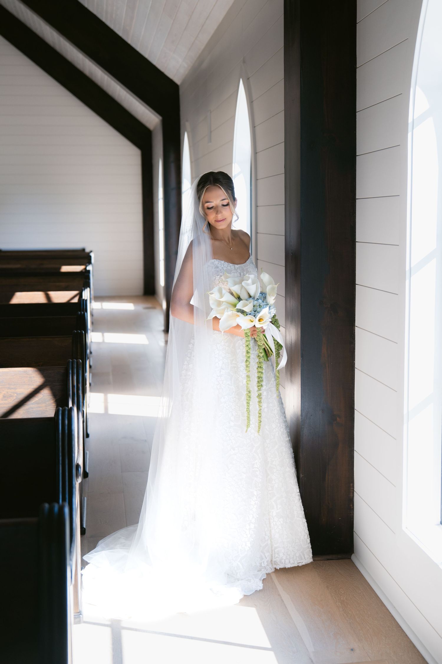 Bride in white lace gown and veil holds floral bouquet by chapel window in soft natural light.